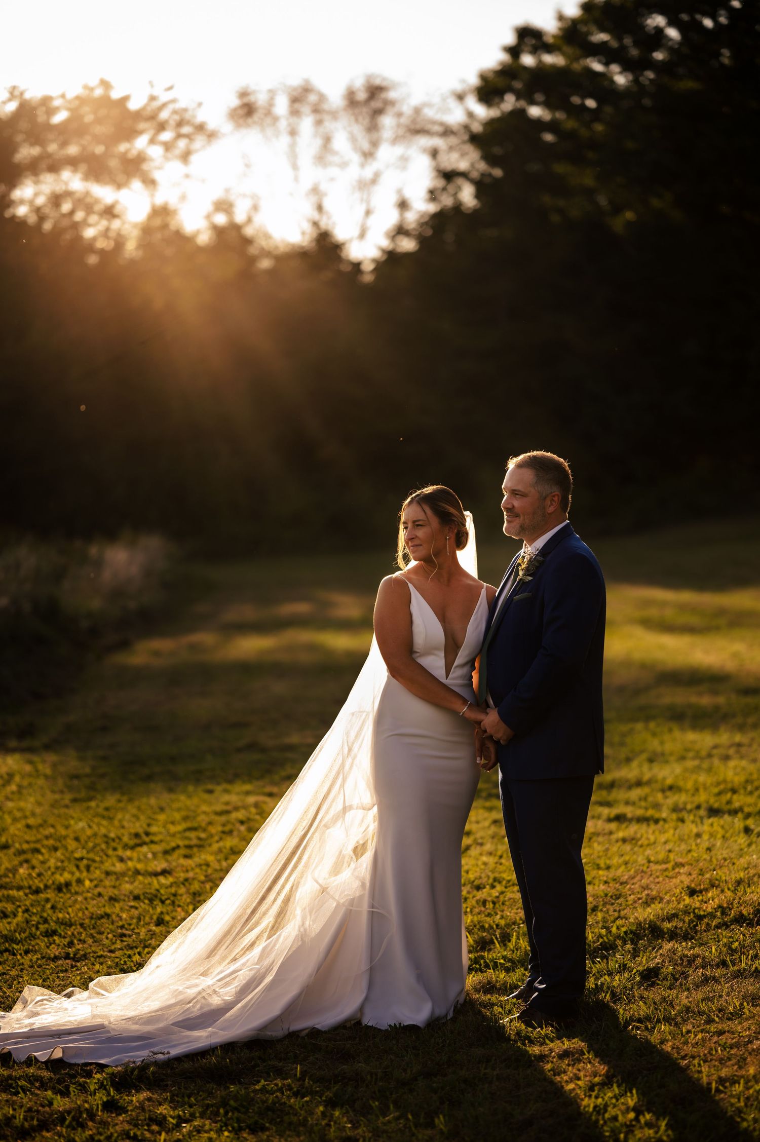 Bride and groom holding hands at golden hour sunset on green grass with warm sunlight rays and trees in background.