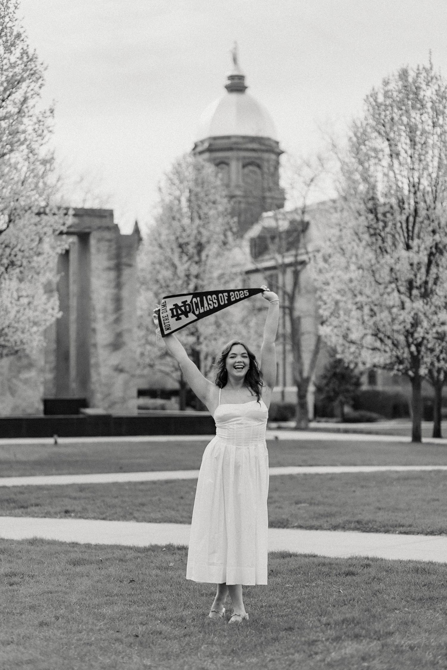 Black and white graduation photo showing a graduate in white dress holding a sign on a campus lawn with domed building behind.