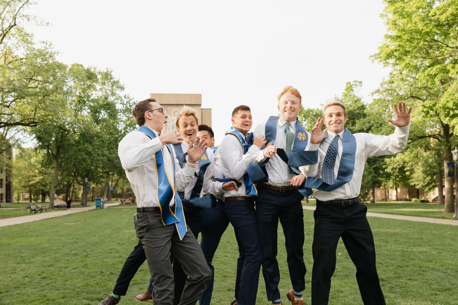 A group of groomsmen in suits lift and celebrate with fun candid expressions on a sunny lawn near trees.