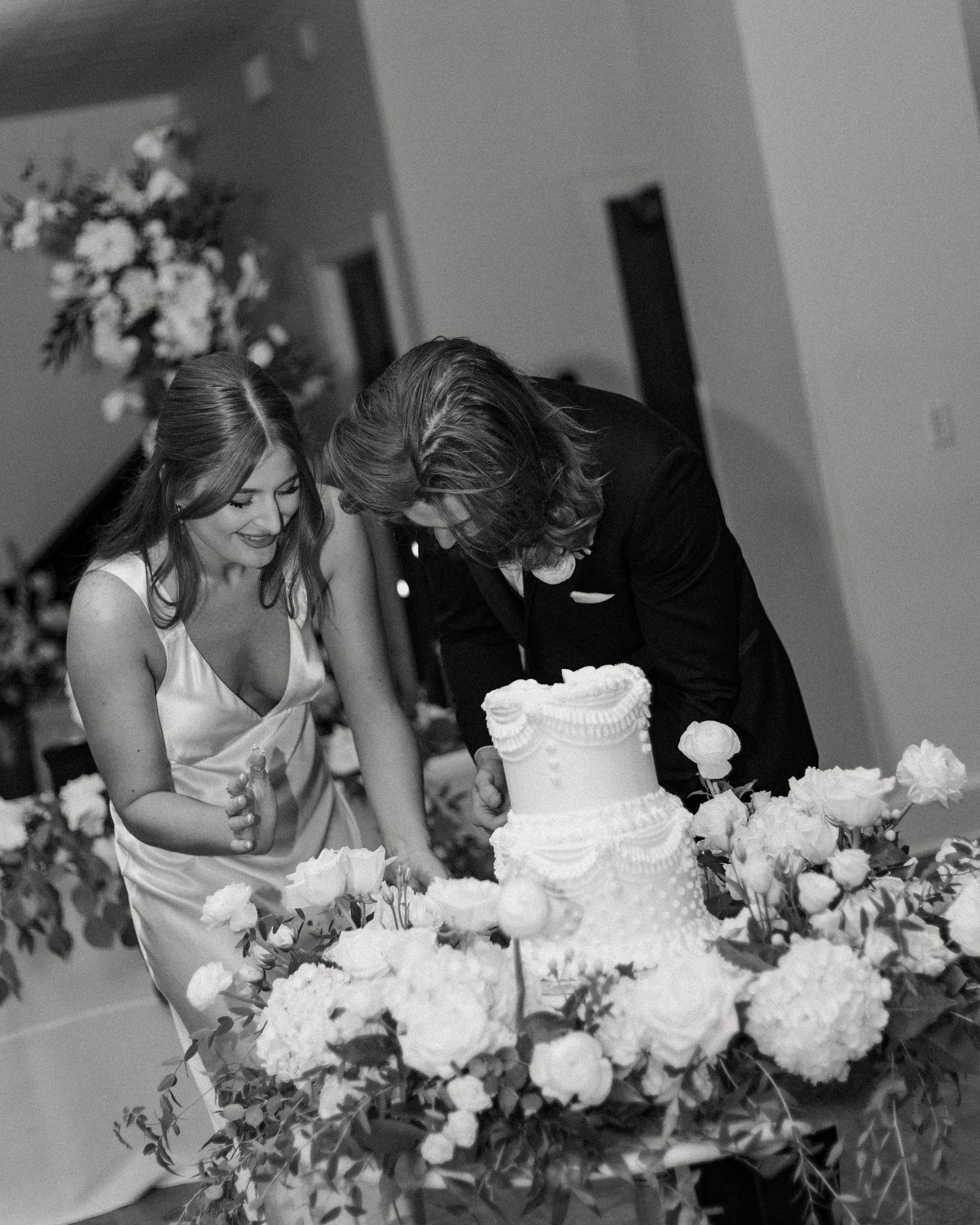 Black and white photo of couple cutting their wedding cake surrounded by white floral arrangements.