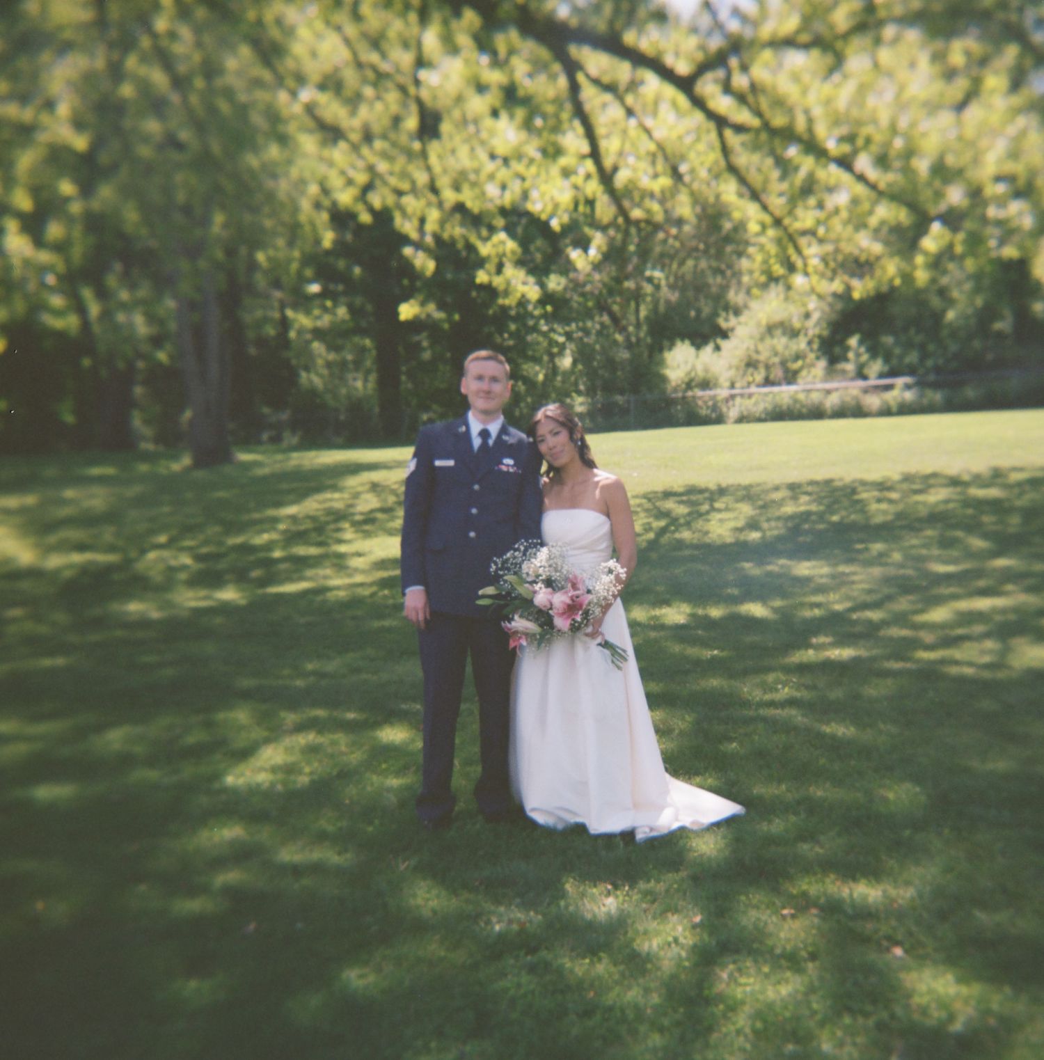 A wedding couple walks and poses together across a sunny green lawn surrounded by trees at an outdoor venue.