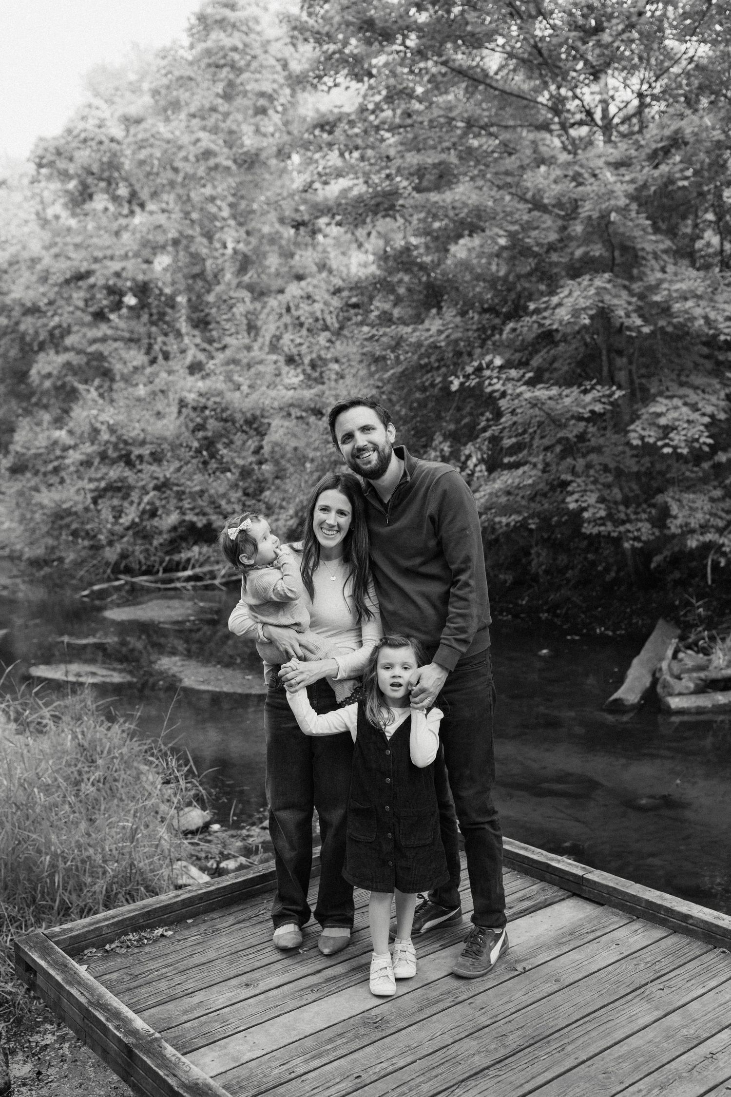 Family of four posing together on a wooden deck by a river.