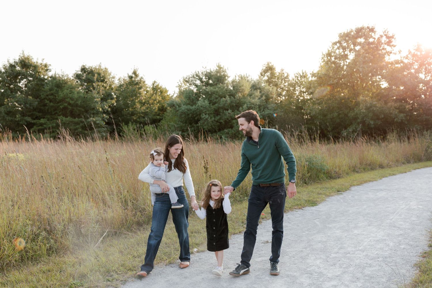 Three people walking together on a paved path through tall grass at sunset.