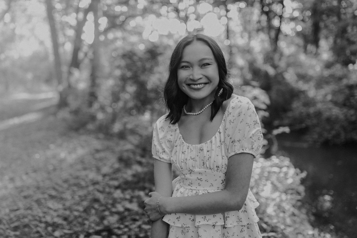 Black and white photo of someone smiling in a floral dress by a stream.