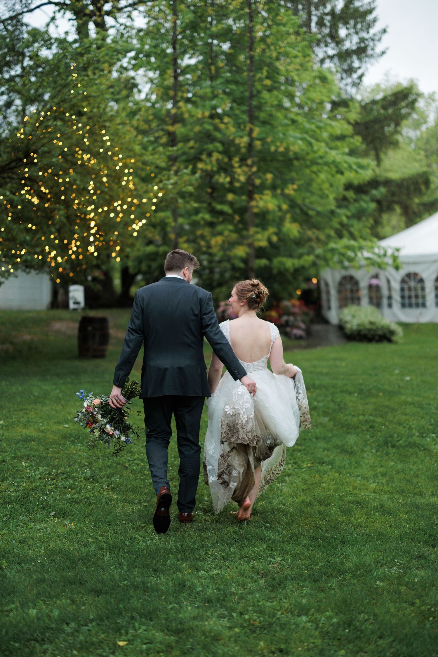 Wedding couple walking across a green lawn with twinkling lights in the background.