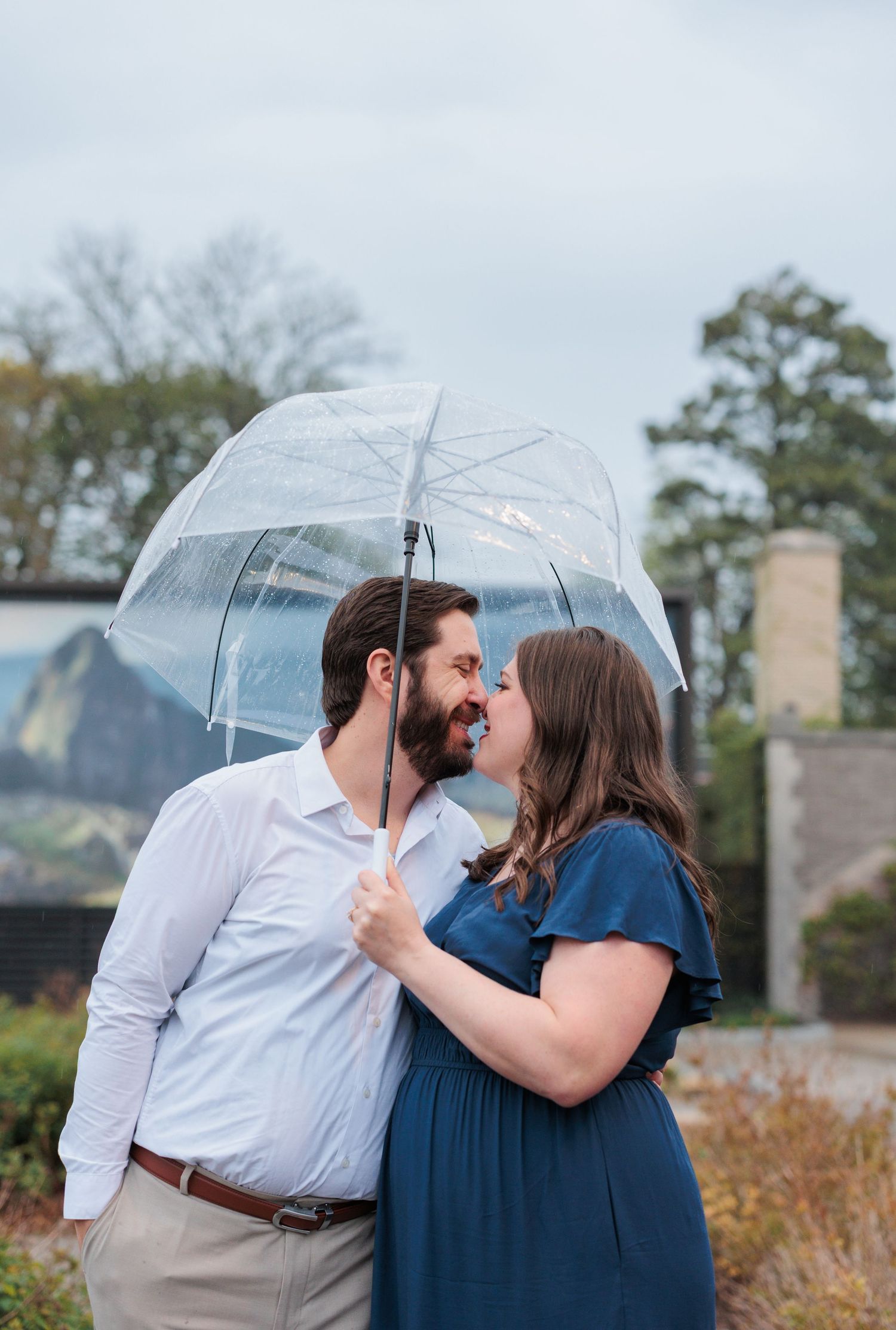 Romantic couple shares intimate moment under clear umbrella during outdoor photoshoot.
