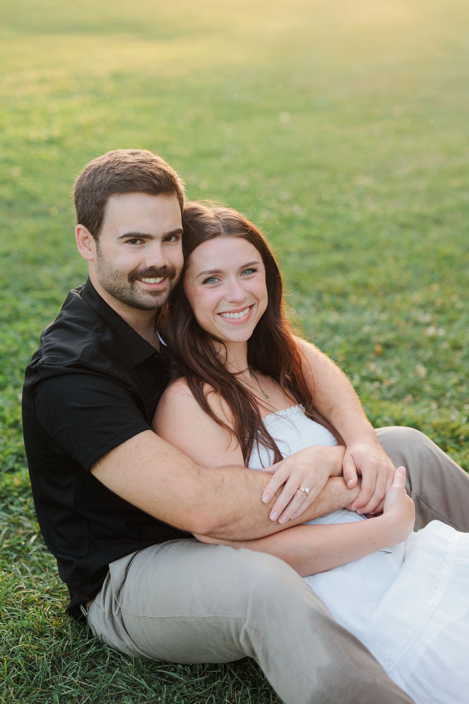 A smiling couple sits on green grass, the man in black embracing the woman in white from behind in warm golden light.