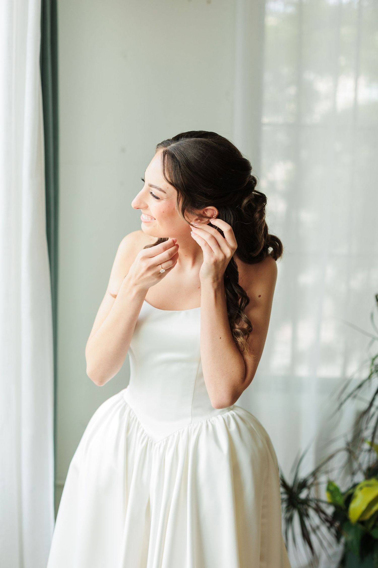 Bride in strapless white gown putting on earrings by a sunlit window, smiling softly on her wedding day.