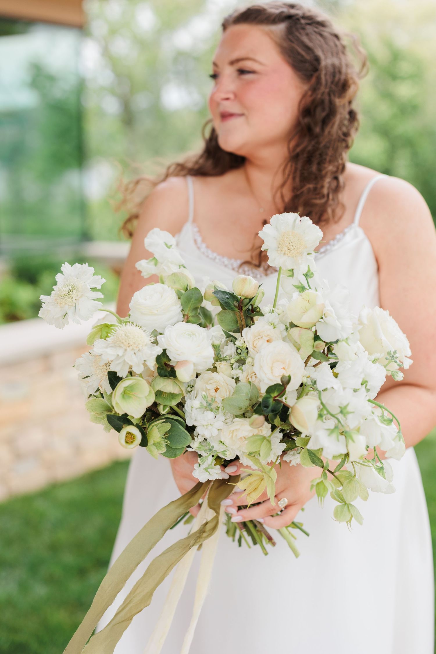 Bride in white spaghetti strap gown holding lush white floral bouquet with greenery and ribbon outdoors.