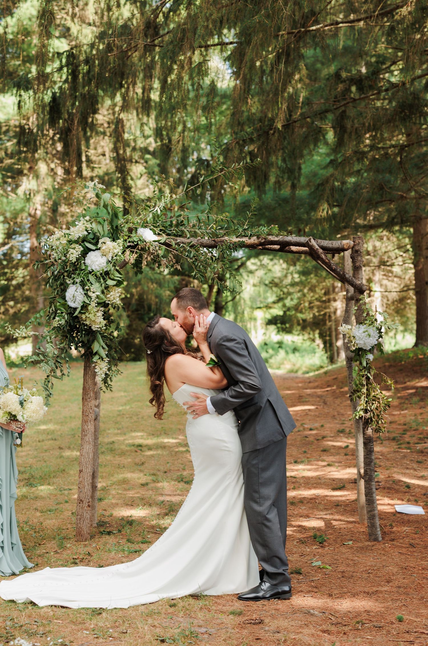 A romantic wedding moment under evergreen trees decorated with hanging floral arrangements.
