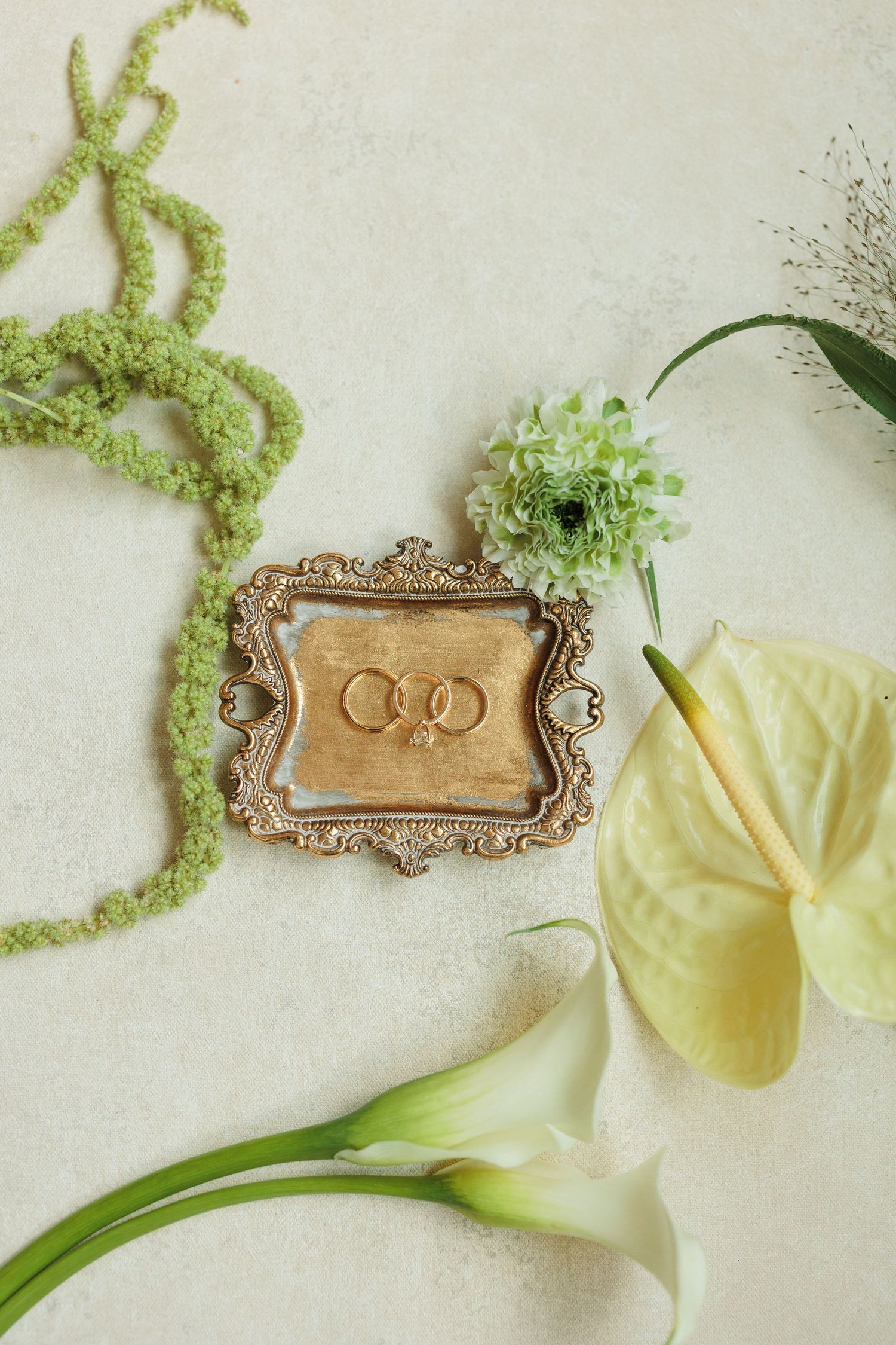 Gold wedding rings in ornate vintage frame surrounded by white calla lilies and green floral accents on neutral background.