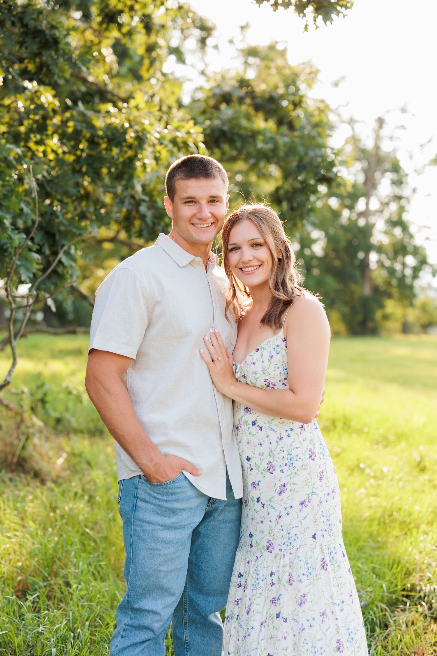 Couple embraces in sunny meadow during summer engagement photoshoot.