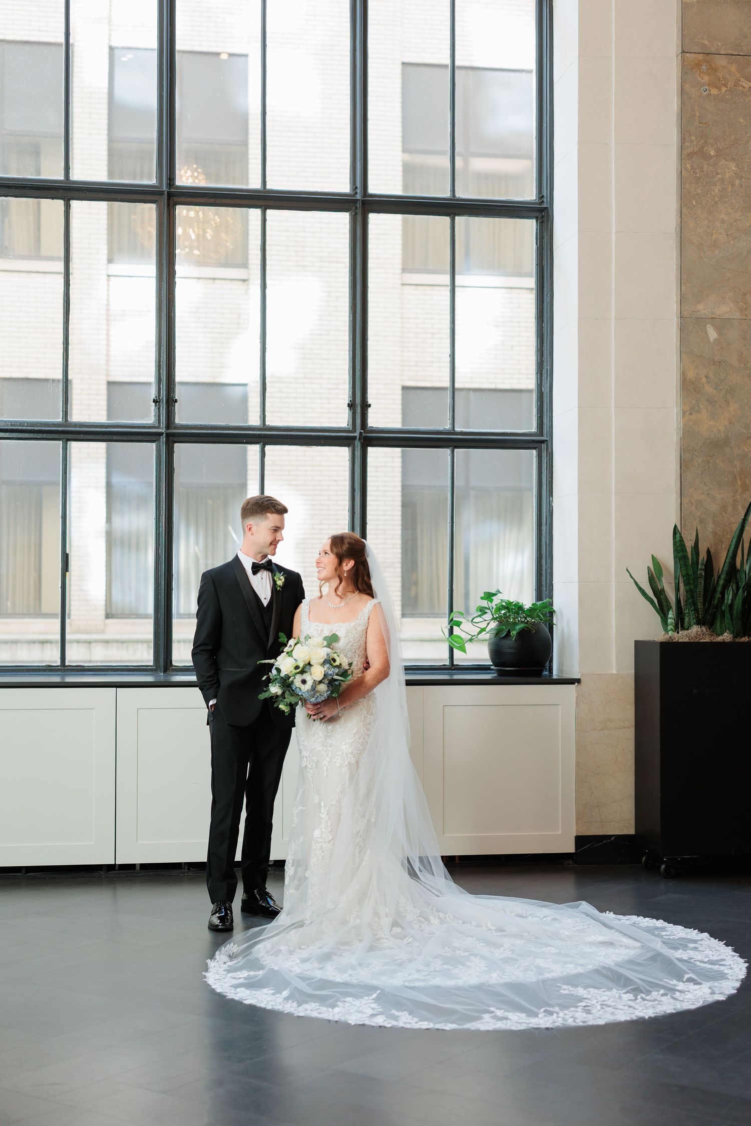 A bride and groom share intimate moments in a modern indoor space with large windows and flowing cathedral veil.