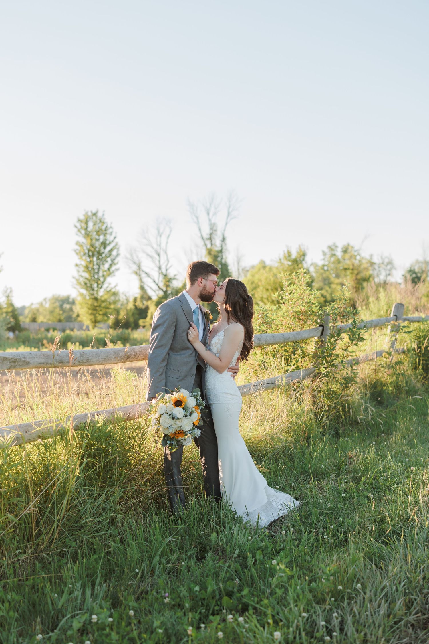A bride and groom share a romantic kiss in a sunlit meadow beside a rustic wooden fence, bride holding a yellow bouquet.