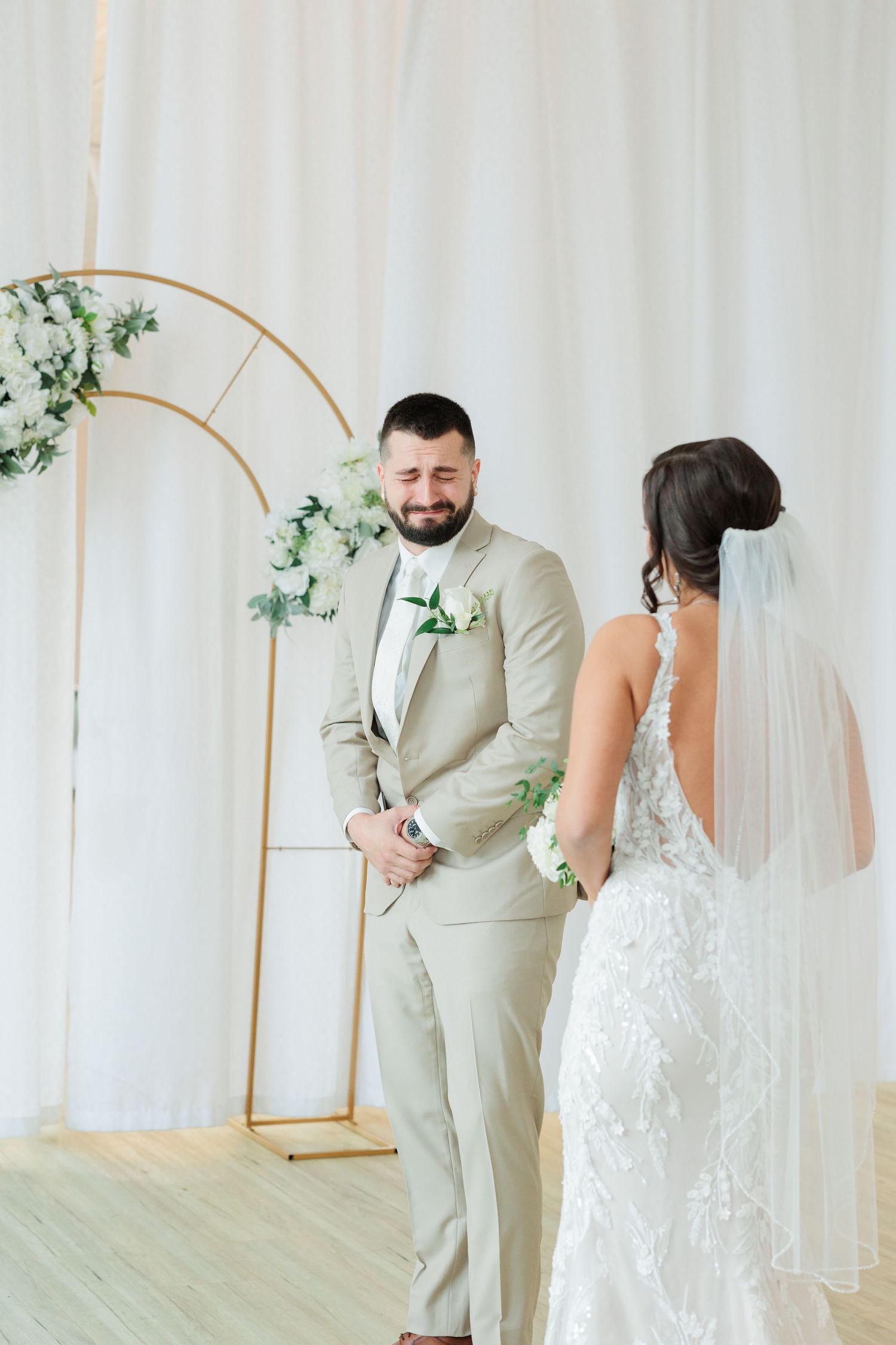 A couple shares their first look moment in a bright studio with white curtains and greenery accents.