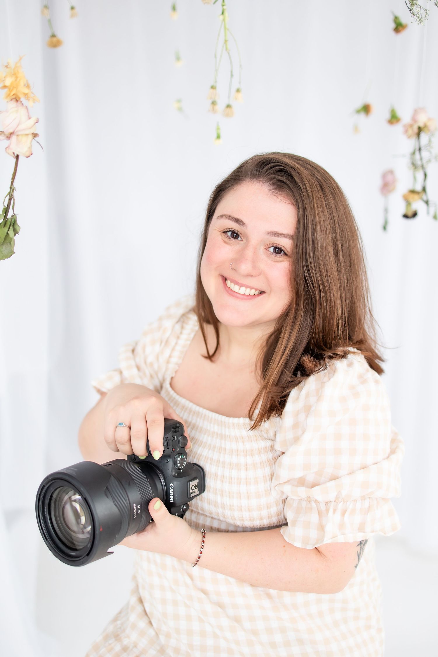 Professional photographer in a peach sweater poses with camera equipment in studio against white backdrop.