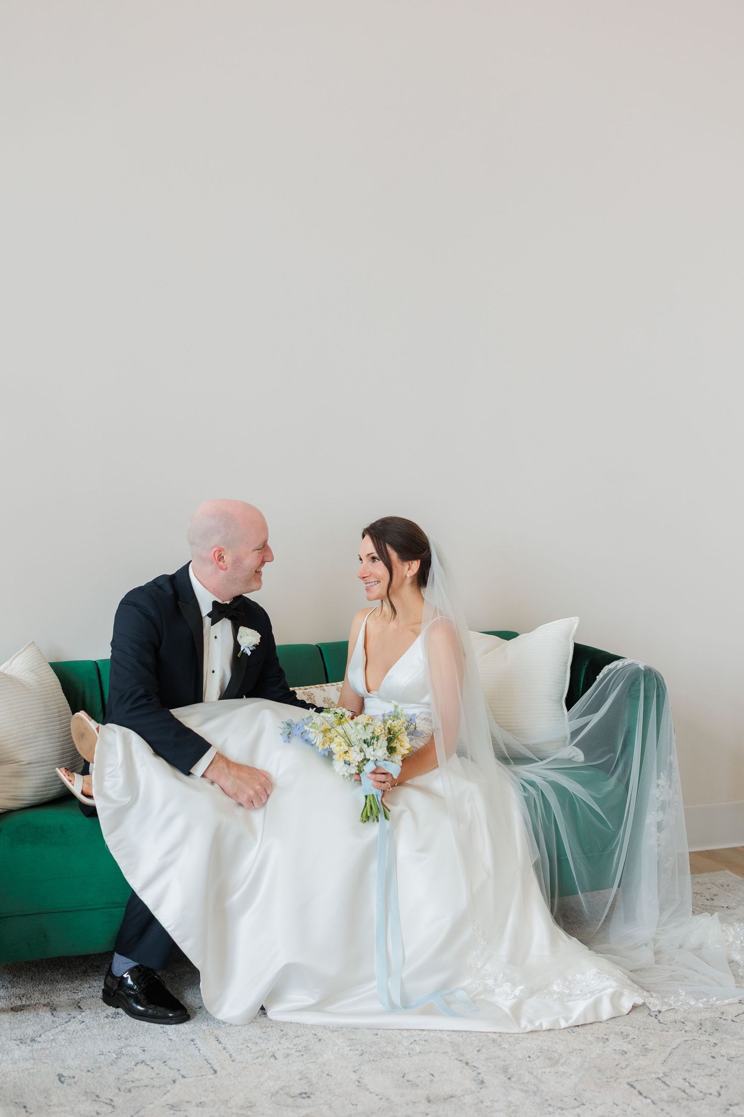 Bride and groom sitting together on a green velvet sofa, smiling at each other, bride holding a pastel bouquet.