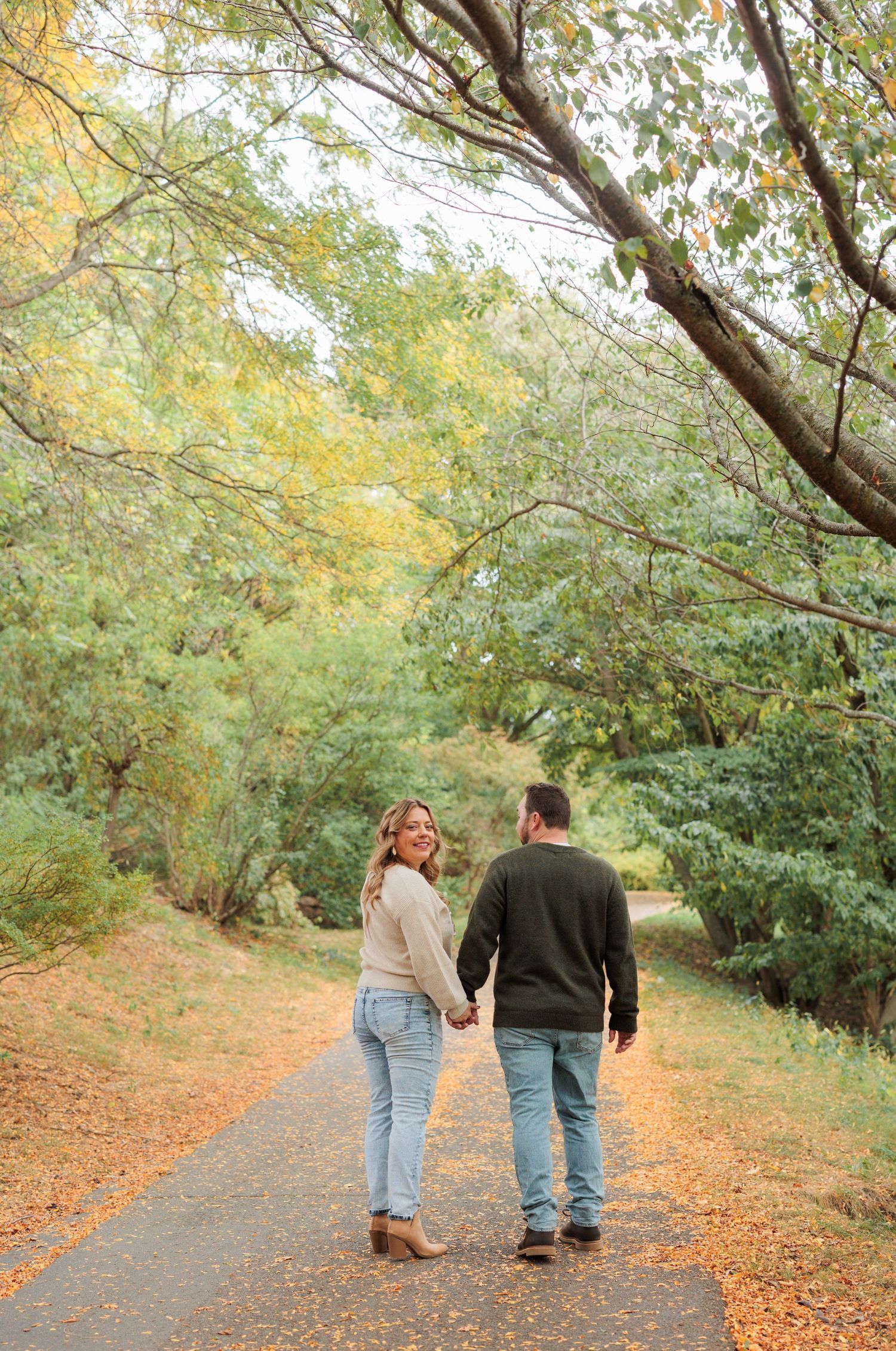 A couple walks together on a leaf-covered path through autumn trees with golden foliage overhead.