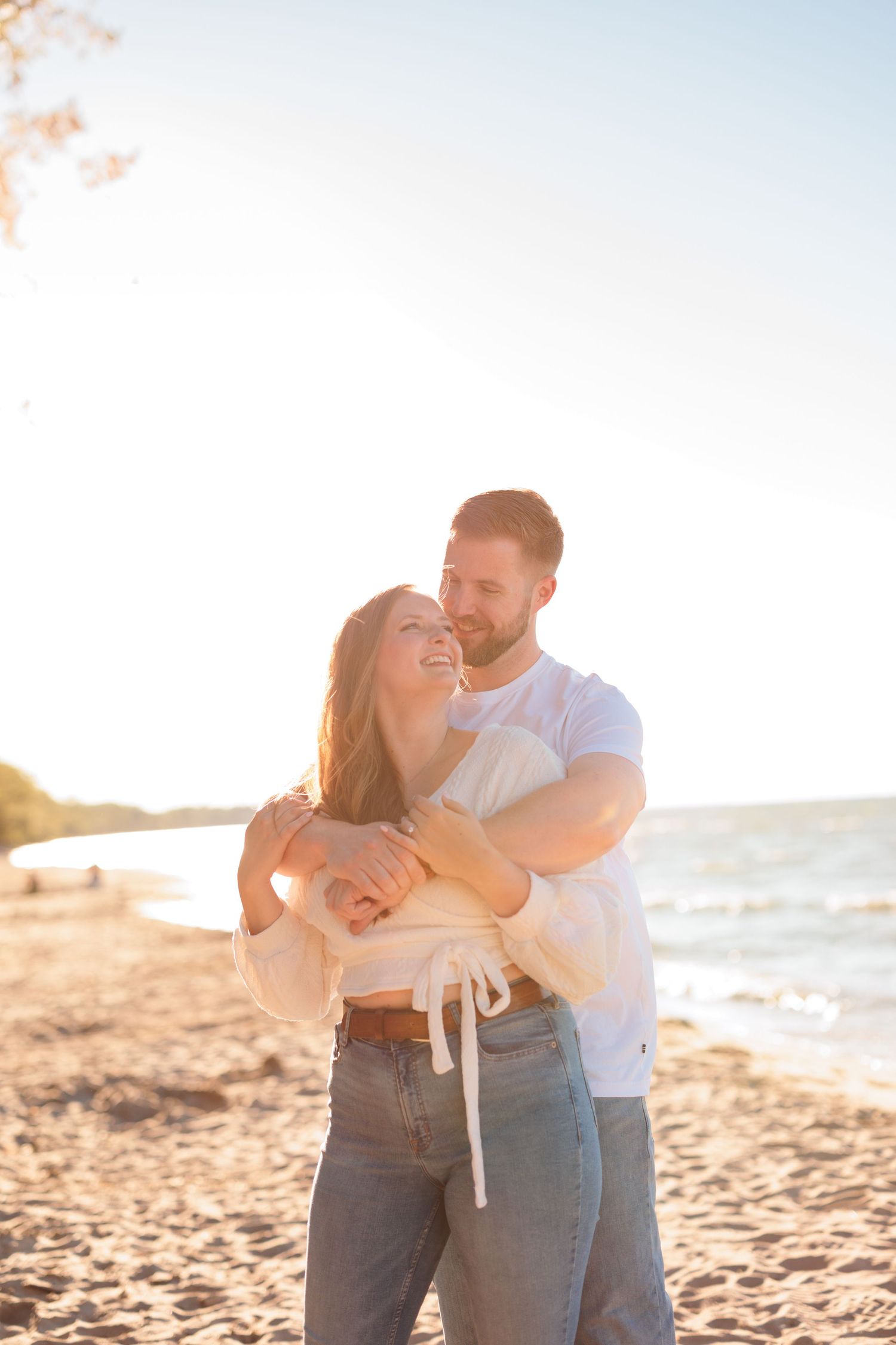 Couple embraces on sunny beach with waves and golden light in background.