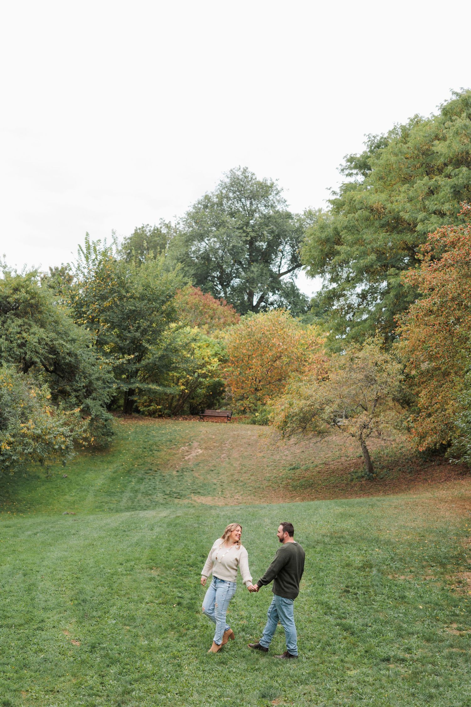 Two people hold hands and dance together on a grassy hillside surrounded by autumn trees in a park setting.
