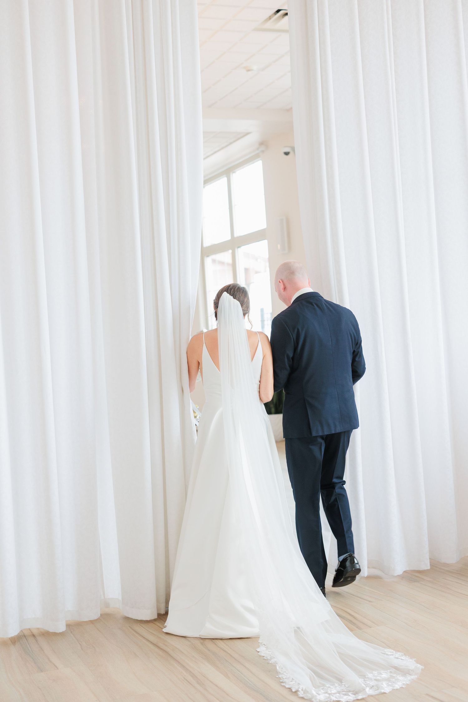 Bride in white gown with veil and groom in navy suit walk toward window through white curtains.