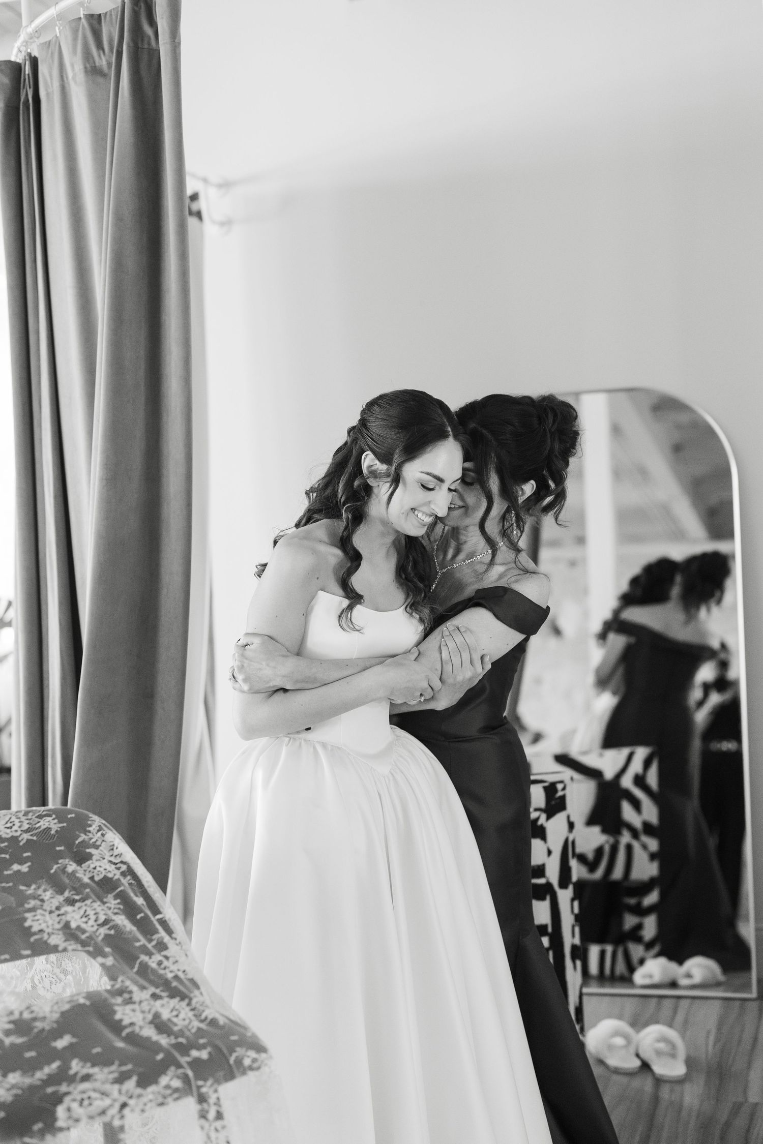 Black and white photo of a bride in a white gown embracing a woman in a dark dress near a mirror.