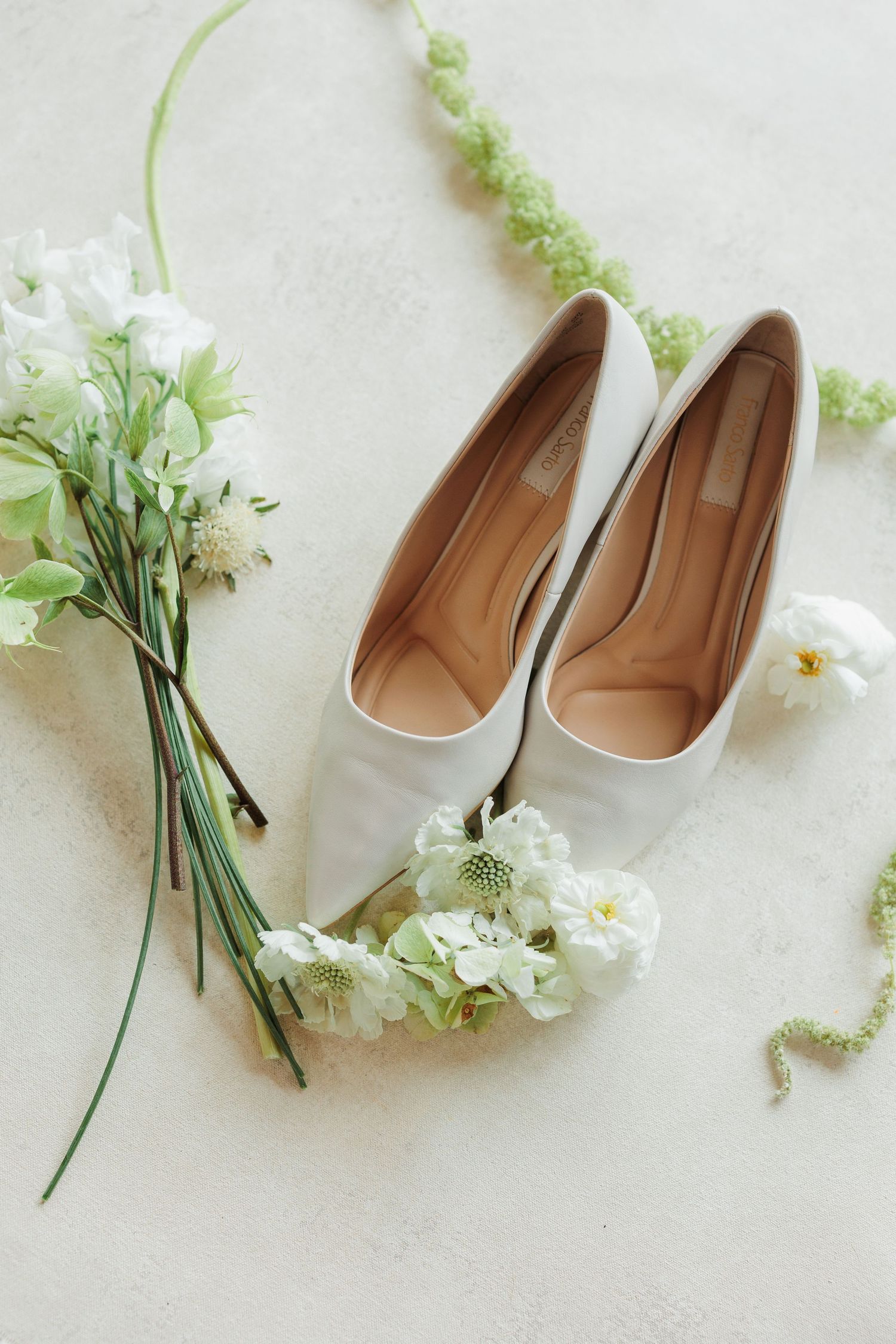 White bridal flats surrounded by delicate white and green flowers on a light surface, perfect for an elegant wedding day.