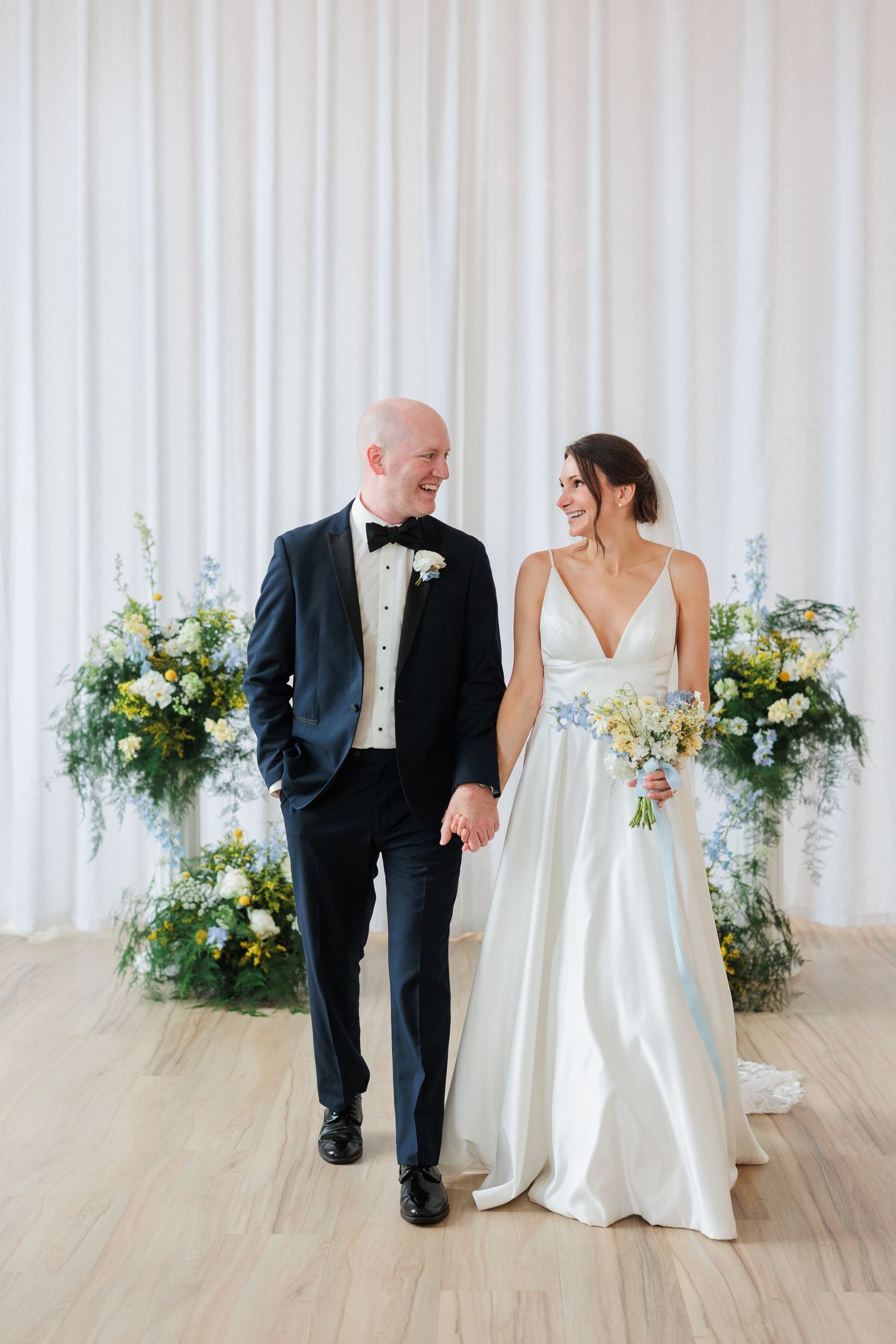 Bride and groom walking hand in hand, smiling, with floral arrangements and white drape backdrop.