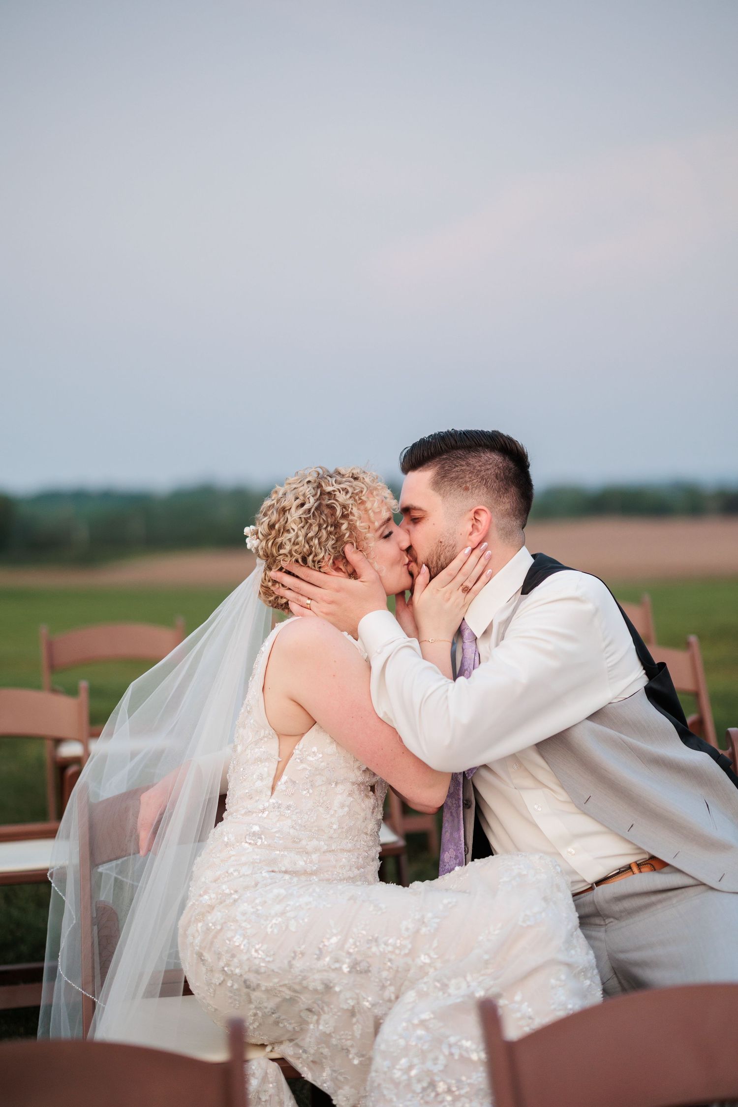A romantic wedding couple shares intimate kisses while seated outdoors during their special day.