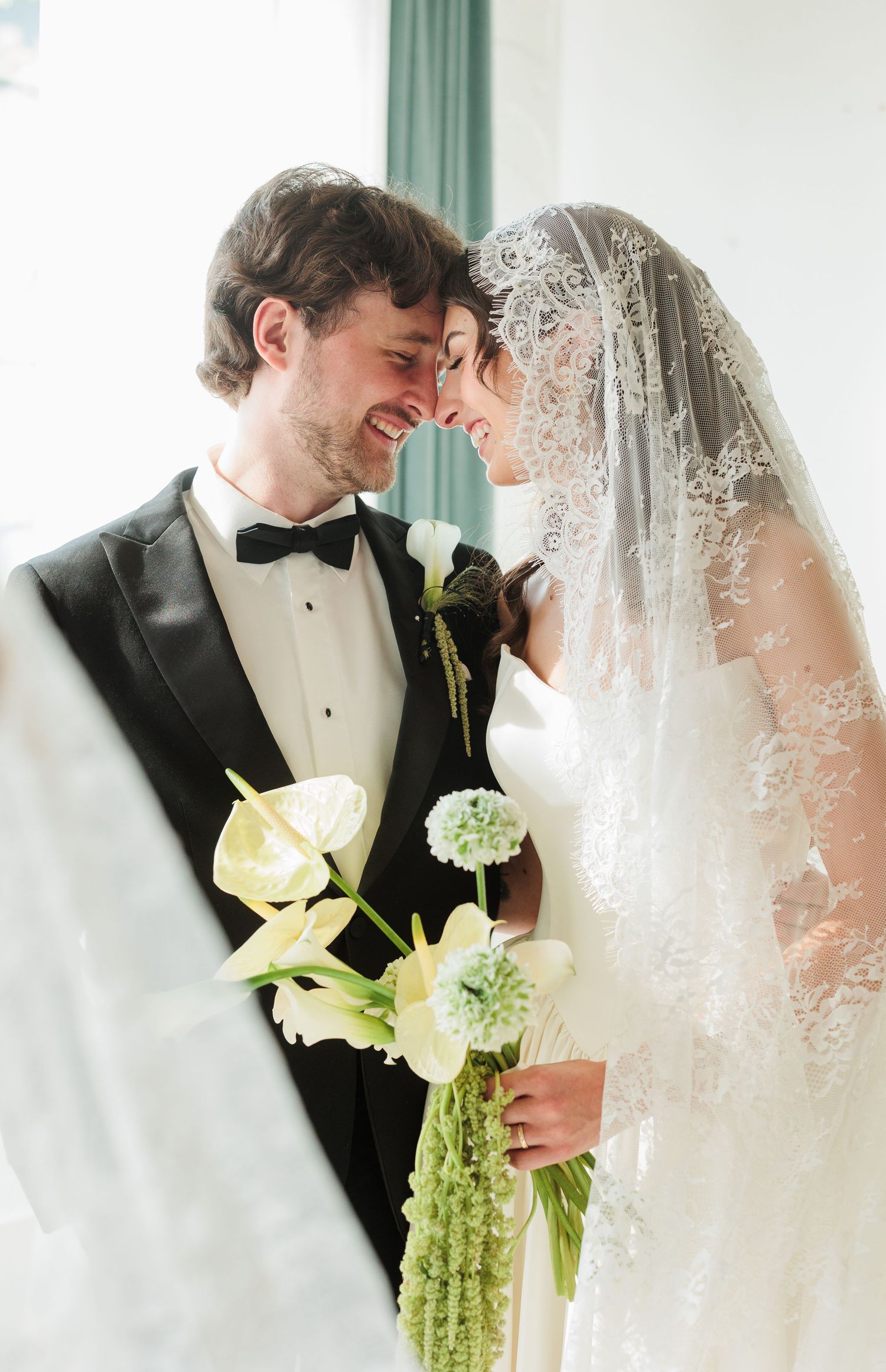 A joyful bride and groom touch foreheads, smiling, as she holds white calla lilies and wears a lace veil.