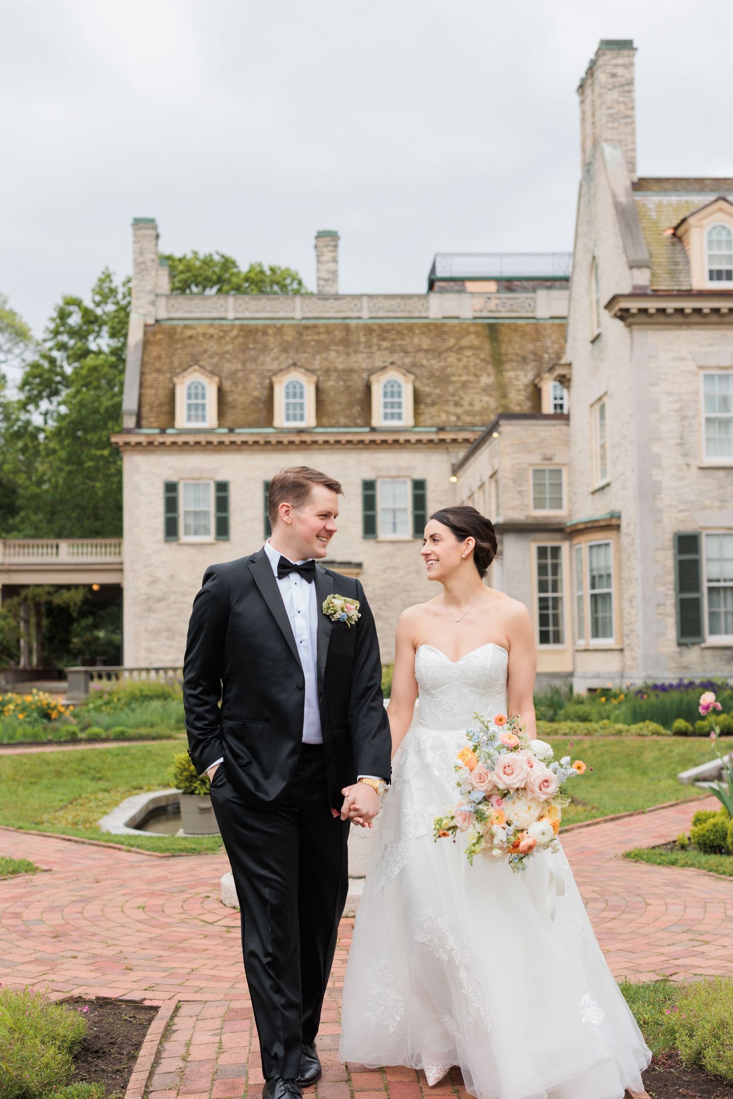 Wedding couple walking together on brick pathway outside historic stone mansion.