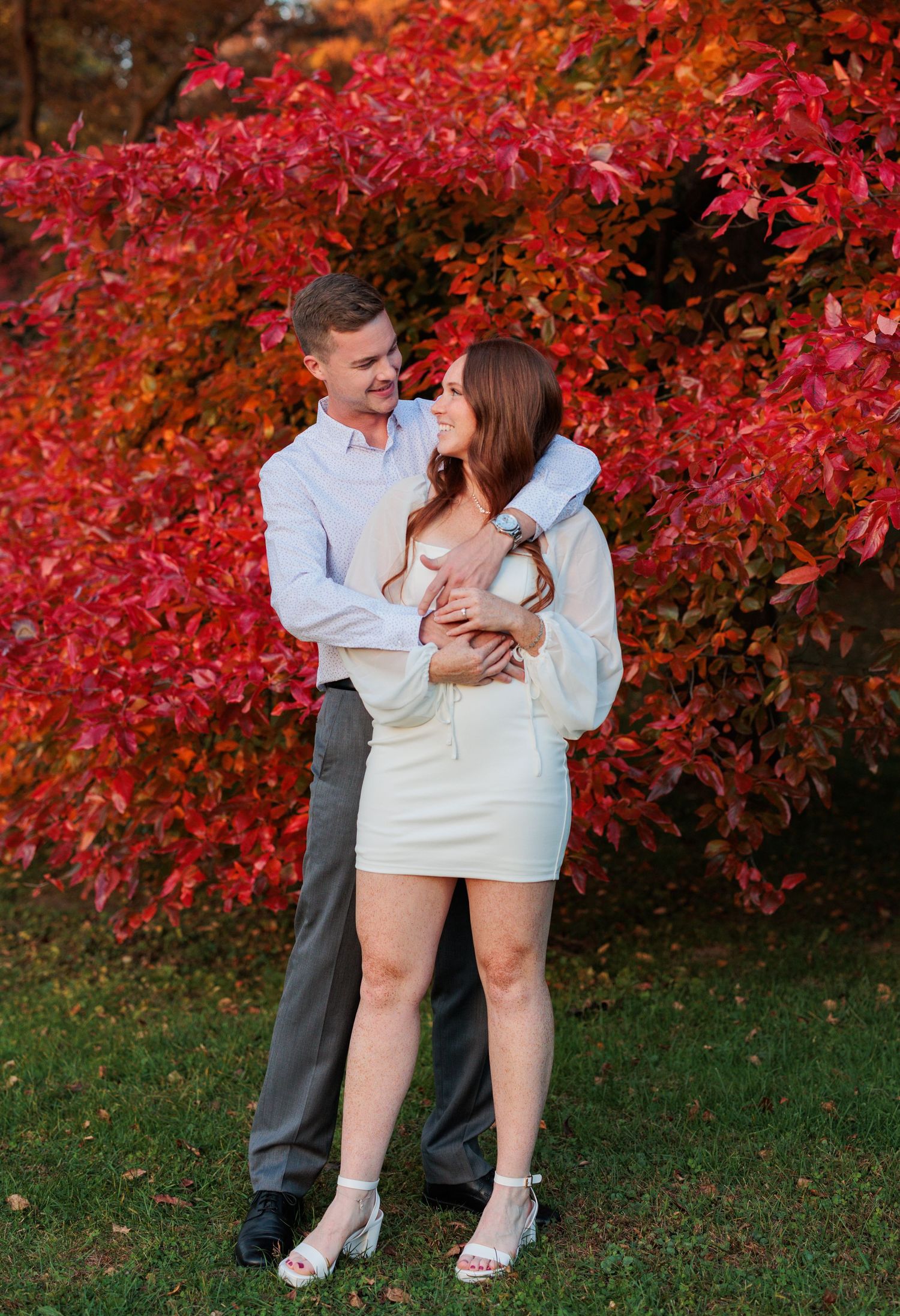 Couple poses playfully against backdrop of bright red autumn bushes during engagement session.