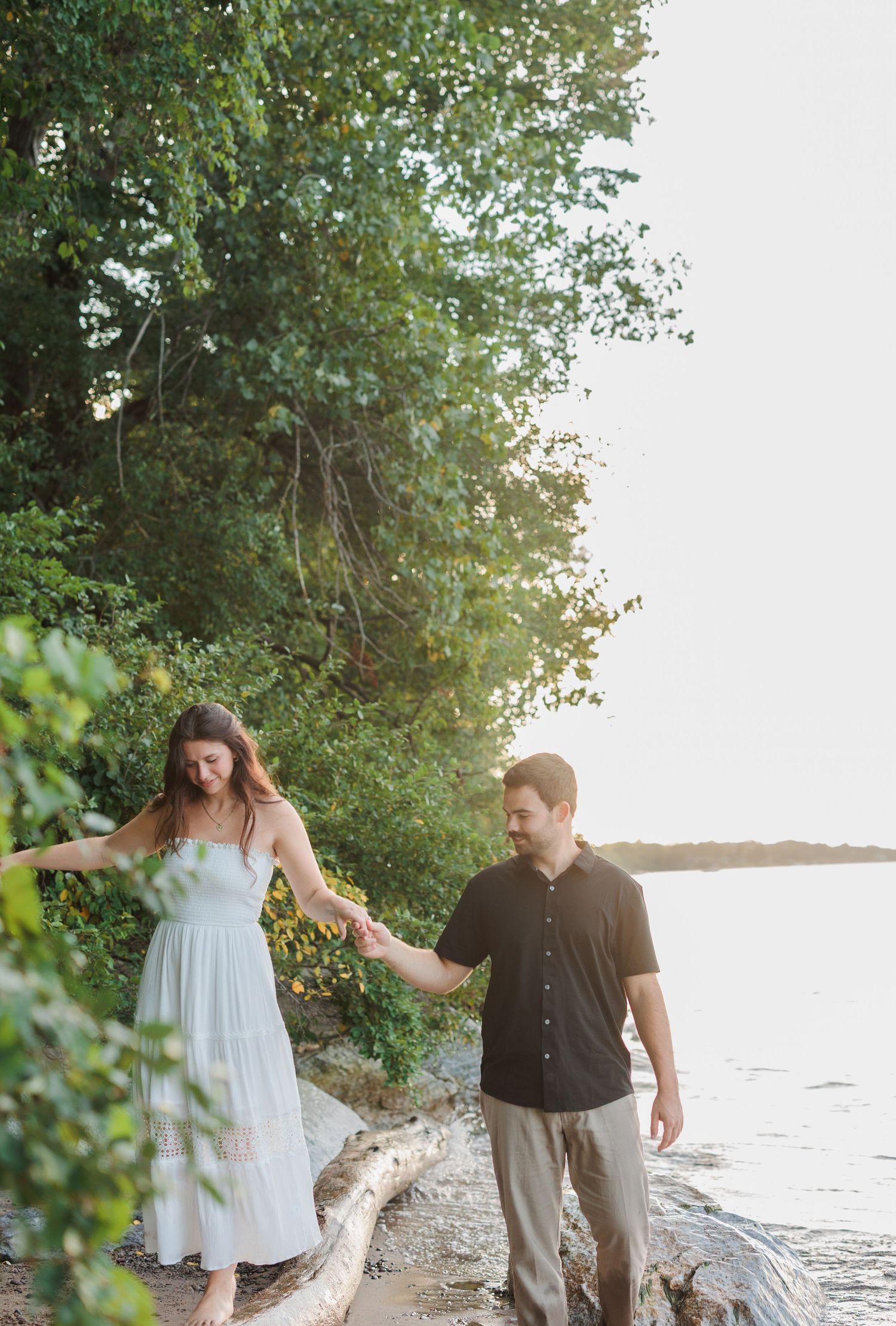 Couple holding hands walking along rocky shoreline surrounded by lush green trees during golden hour.