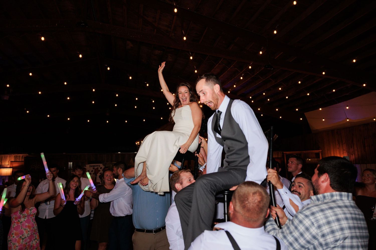 Wedding reception guests dance and celebrate under twinkling lights in a dark venue.