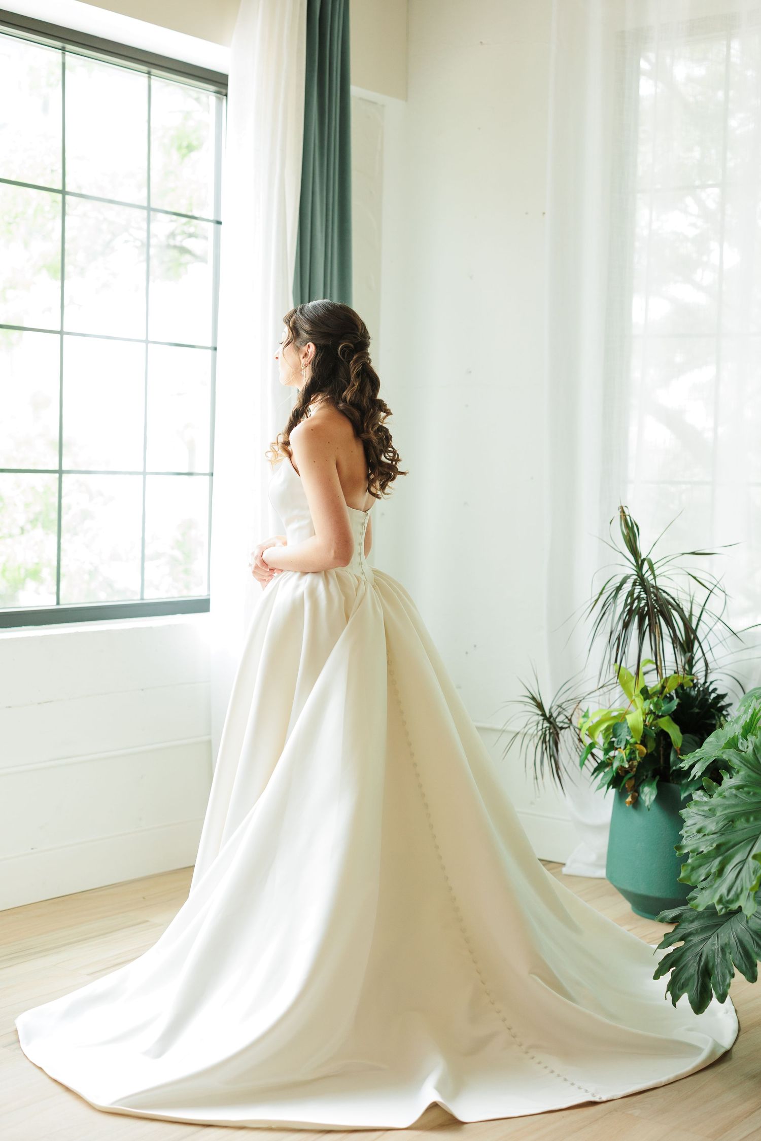Bride in elegant white ball gown with long train stands by large window, looking out with lush green plants nearby.