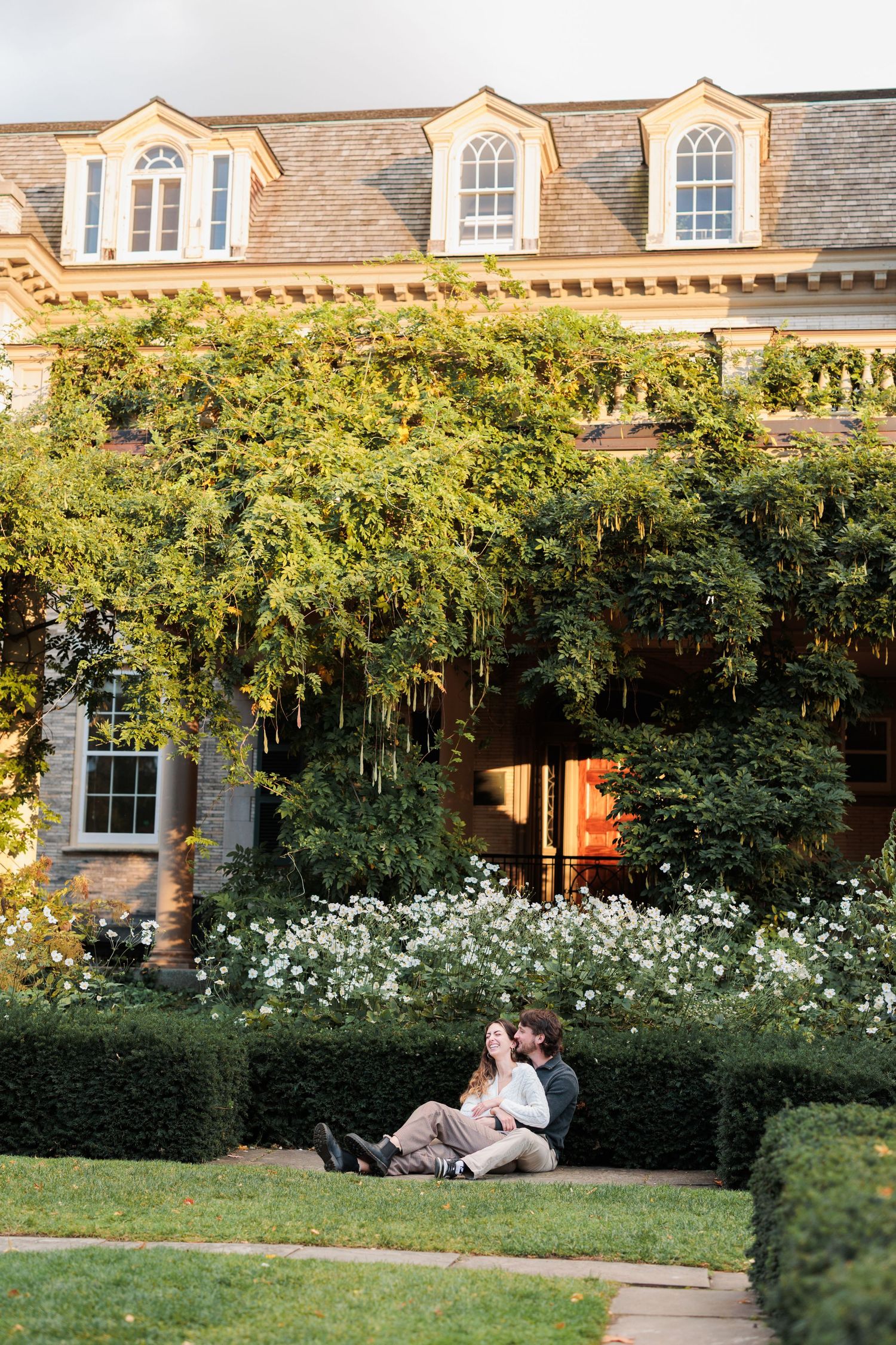 A couple sits together on a garden bench in front of a stone building with climbing vines and white flowers.