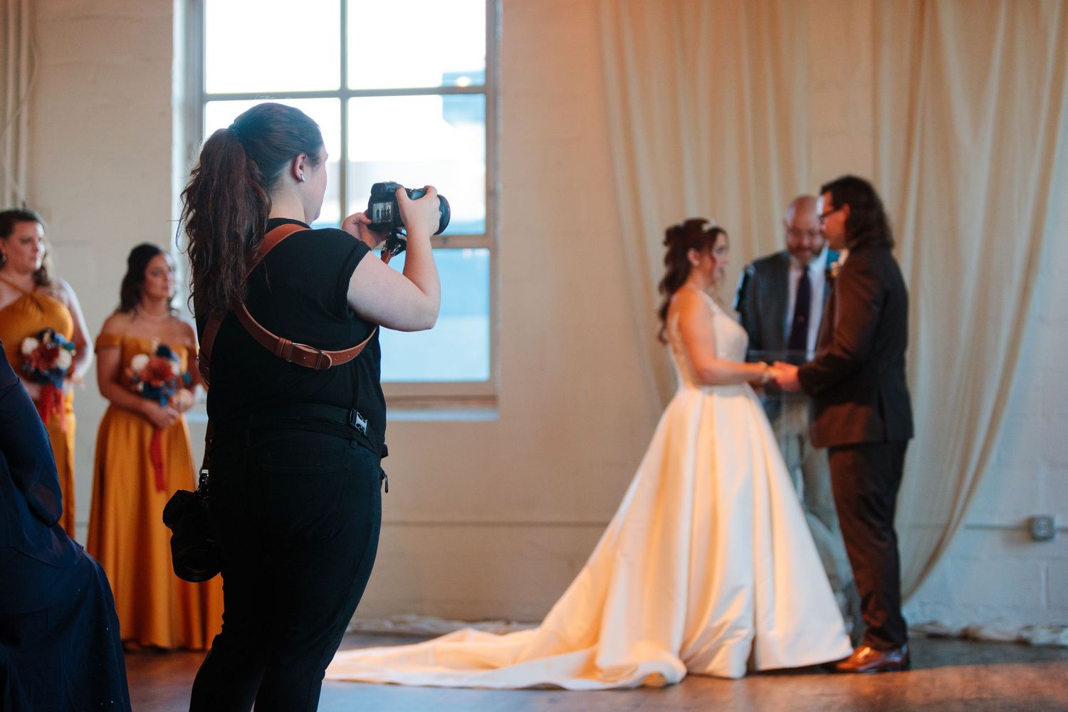 Wedding party gathered in an indoor venue with white curtains and natural lighting.