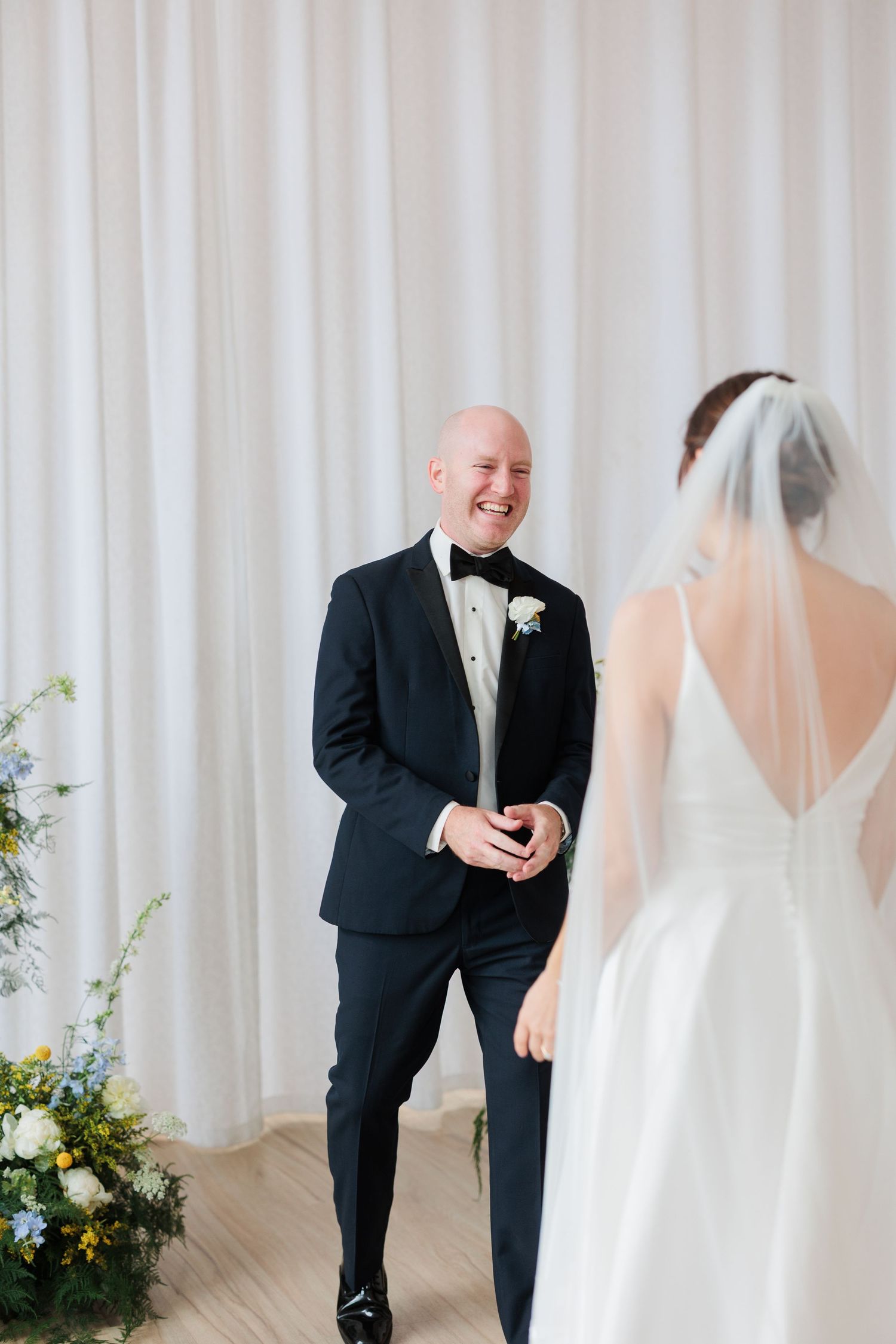 Groom in black tuxedo smiling joyfully at bride in white gown during wedding first look ceremony.