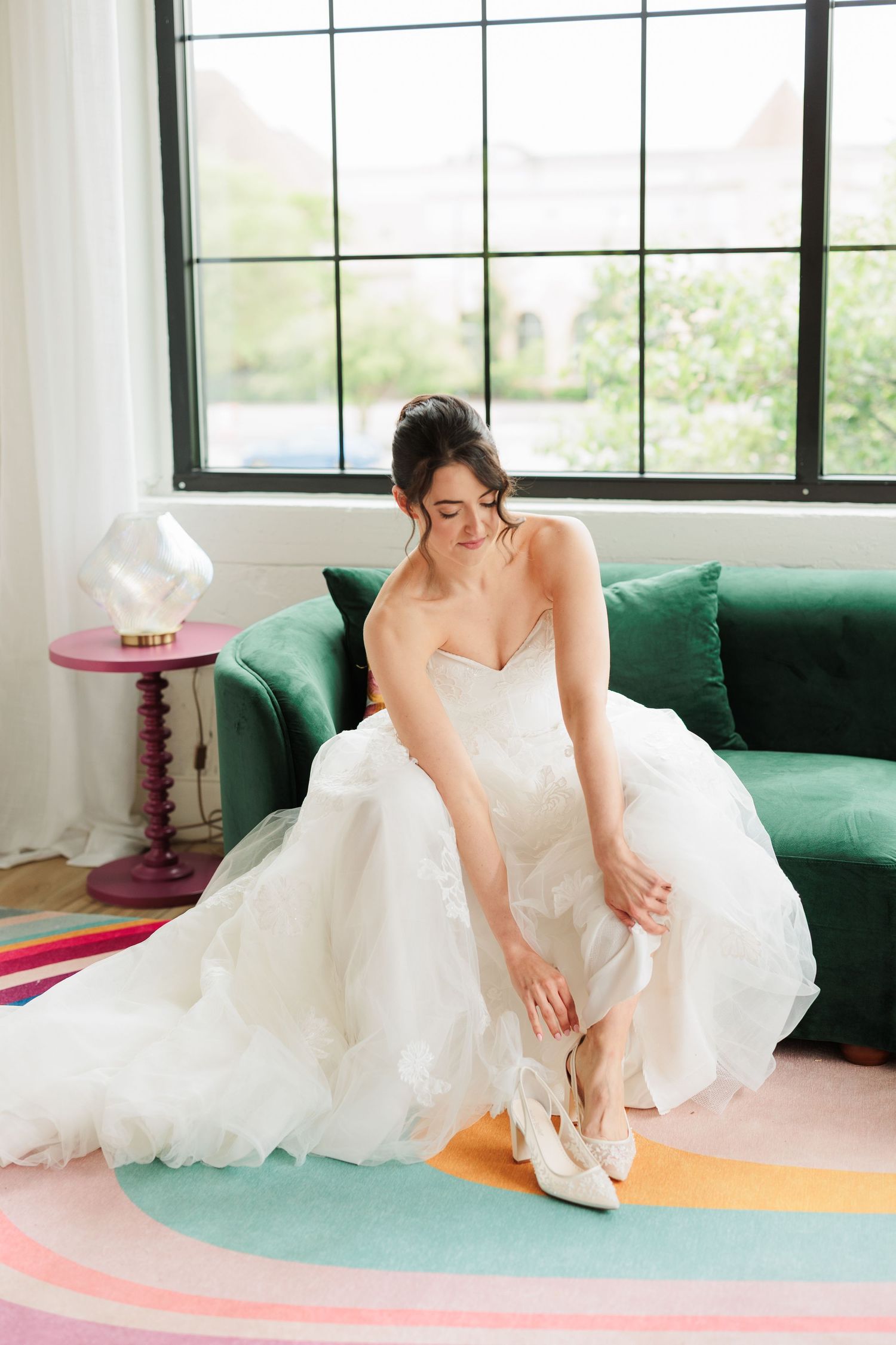 A bride in a flowing white gown seated on a colorful floor near an emerald green couch.