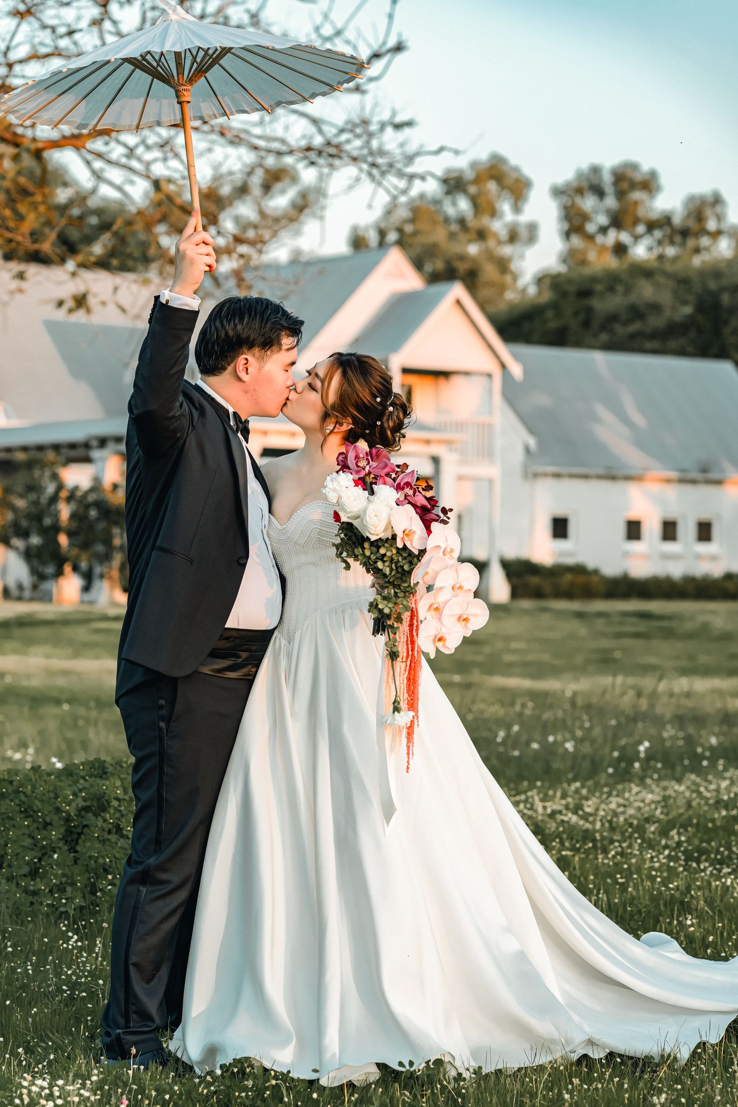 A romantic wedding couple shares a kiss in a garden setting while holding a delicate white parasol against a rustic barn backdrop.