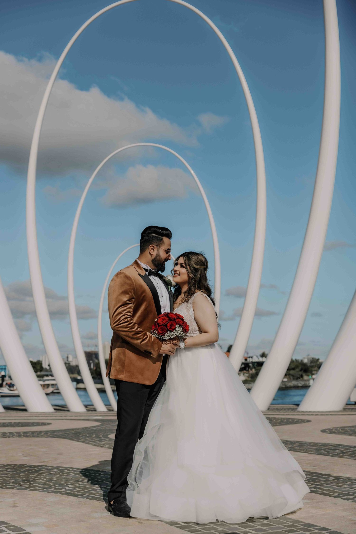 A wedding couple embraces under white architectural arches against a blue sky backdrop.