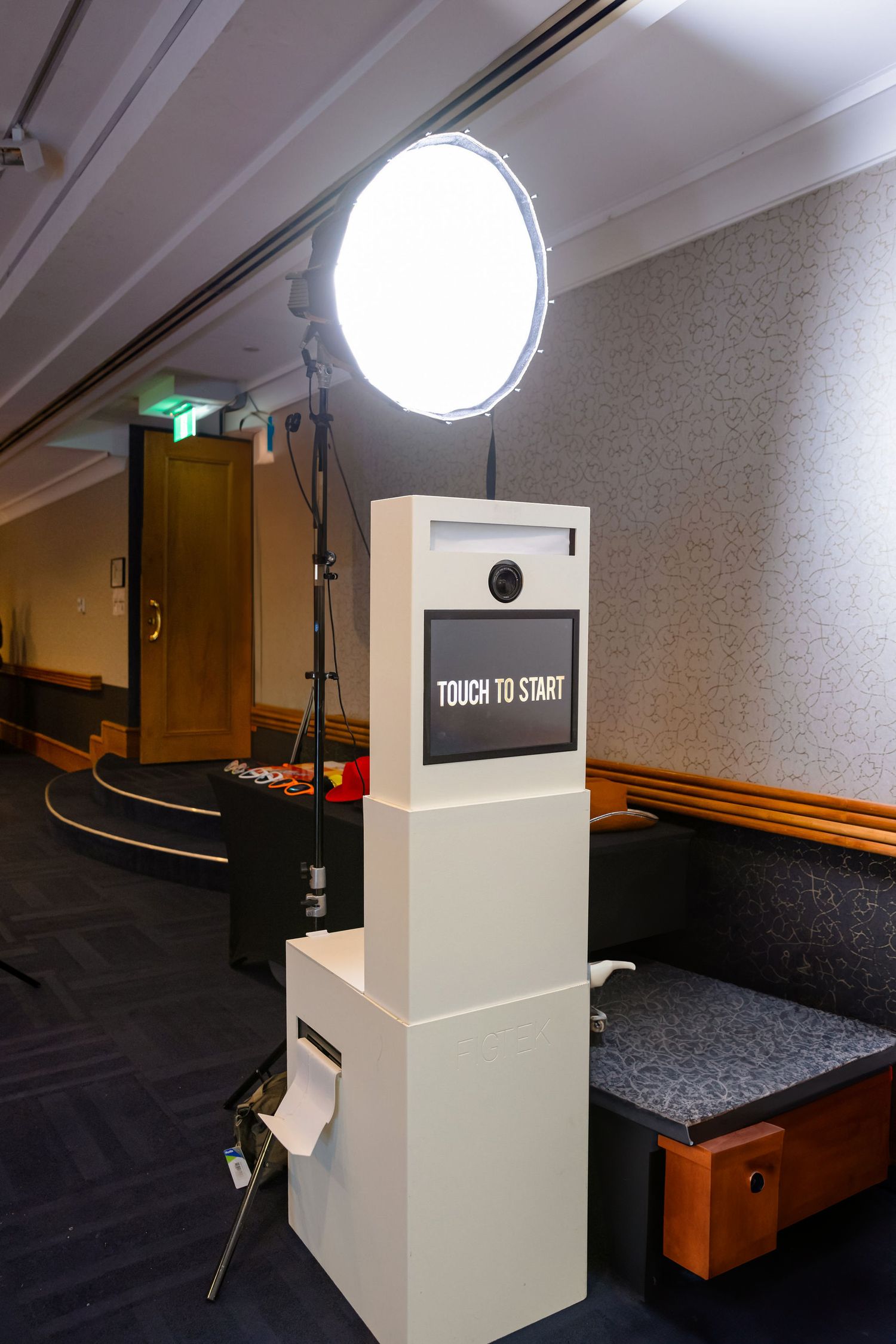 A photo booth setup with a bright circular light and 'Touch to Start' display screen mounted on a white kiosk stand.