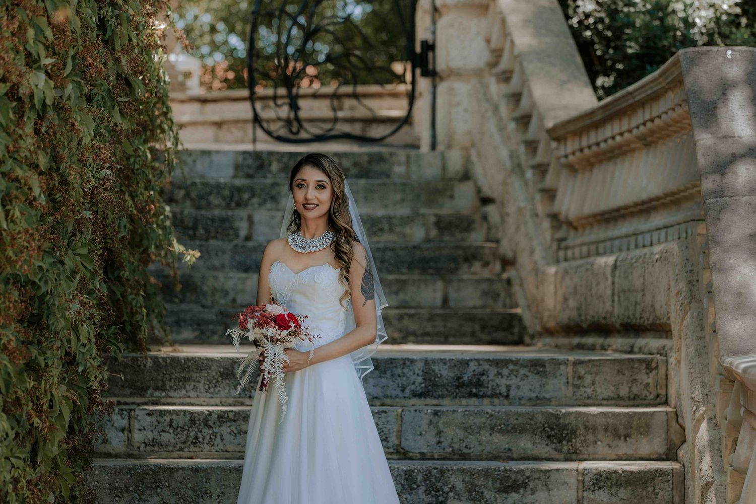 A bride in a white gown holds a bouquet while standing on ornate stone steps.
