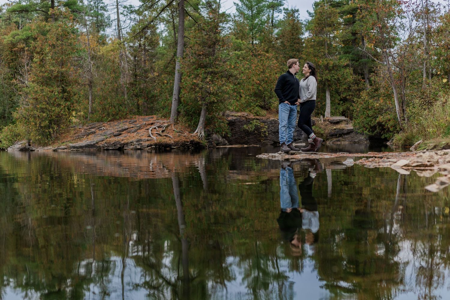 Relaxed engagement photo of couple standing together by the water surrounded by trees at the rockwood conservation area.