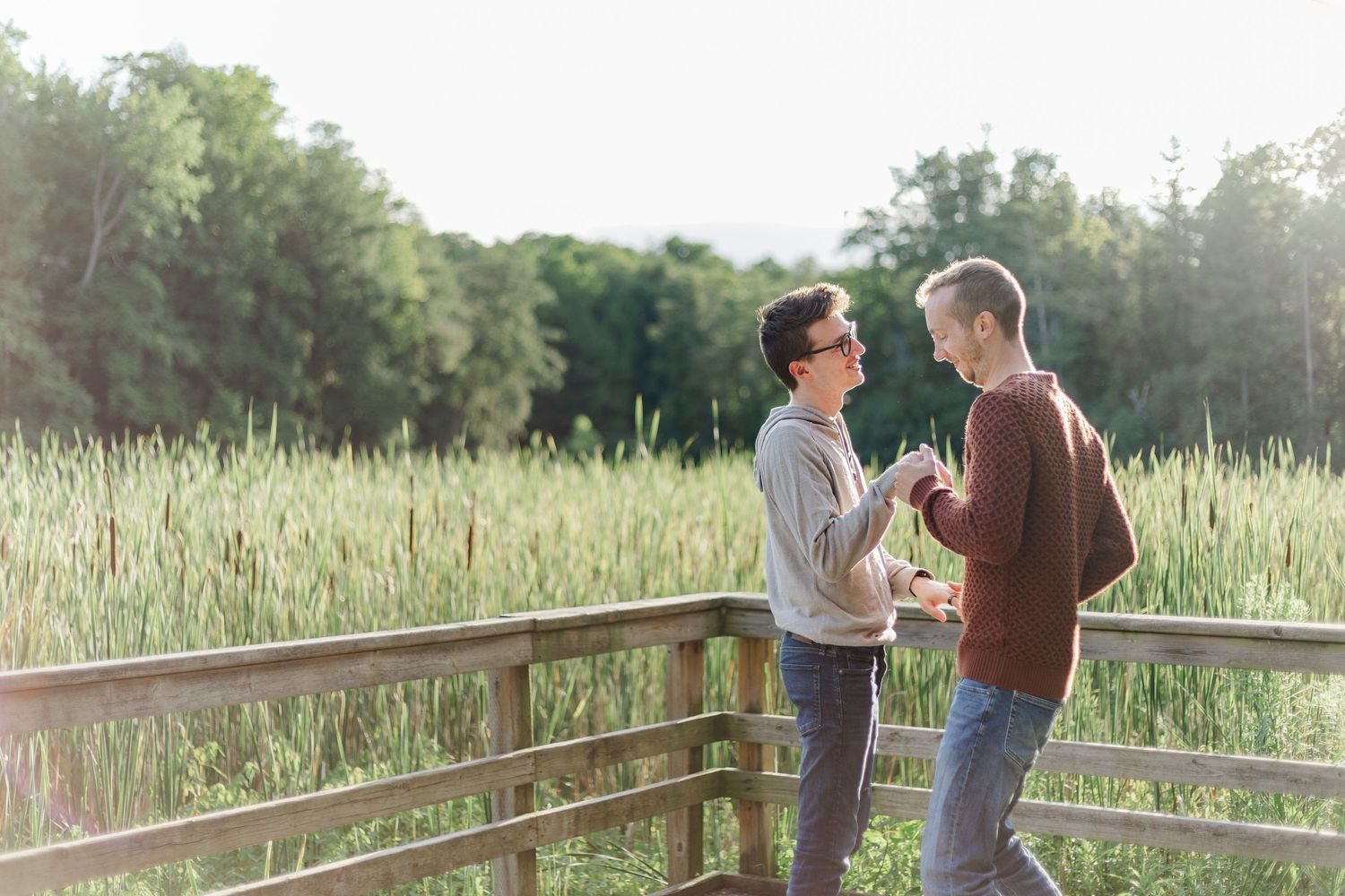 A couple stands together in a sunlit meadow with tall grass and wildflowers during golden hour.
