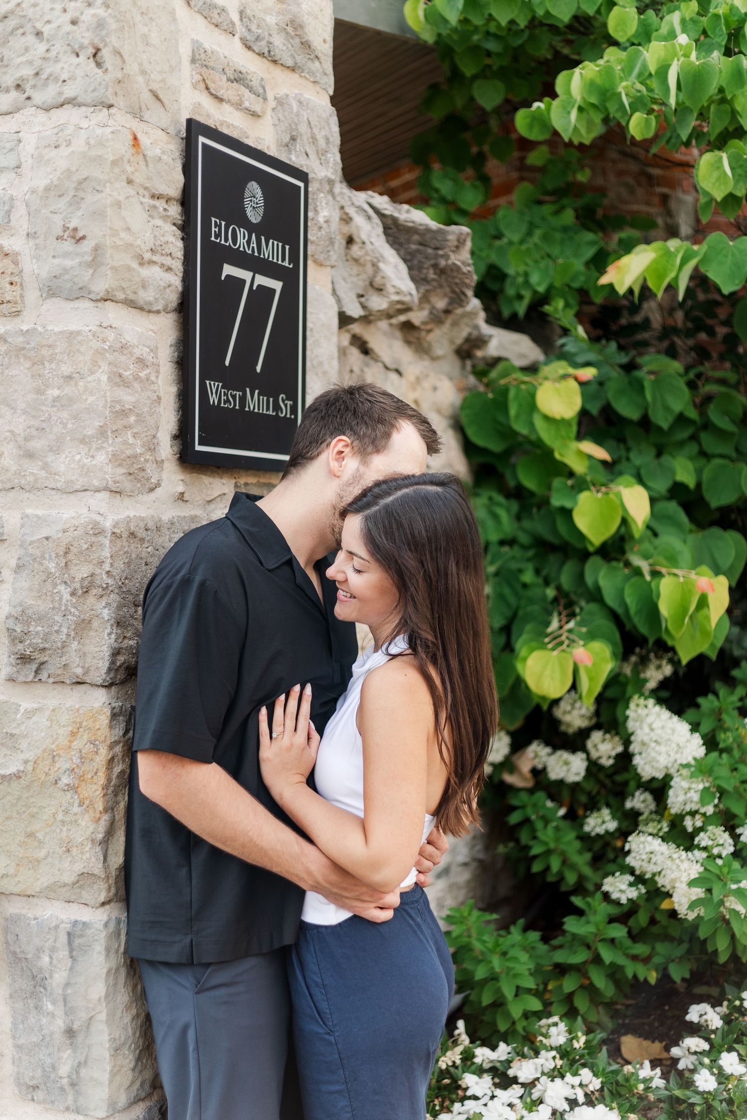 A couple shares an intimate moment against a stone wall with green foliage and white flowers in the background.