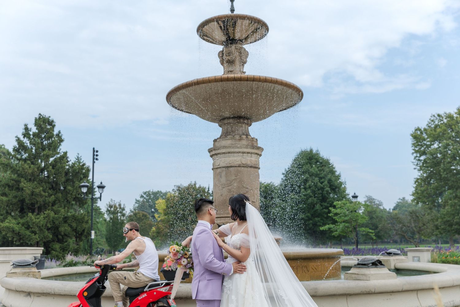 Candid wedding photo of bride and groom watching a moped drive by in front of the fountain at gage park in hamilton, ontario.