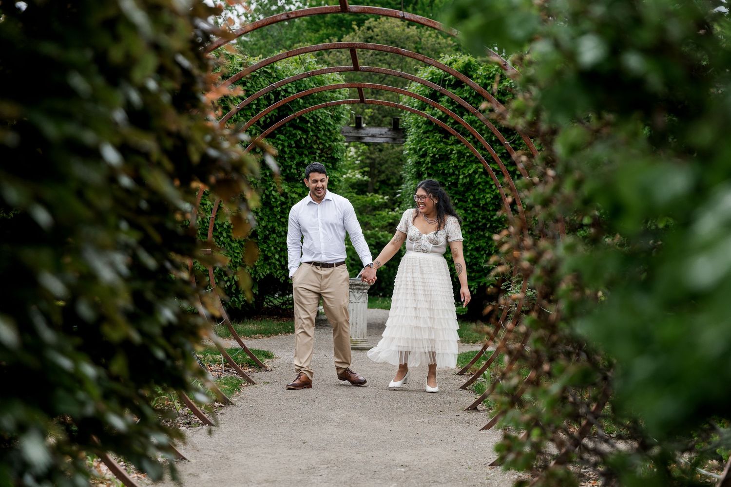 A couple walks along a garden path surrounded by lush greenery and foliage during a romantic engagement session at The Guelph Arboretum.