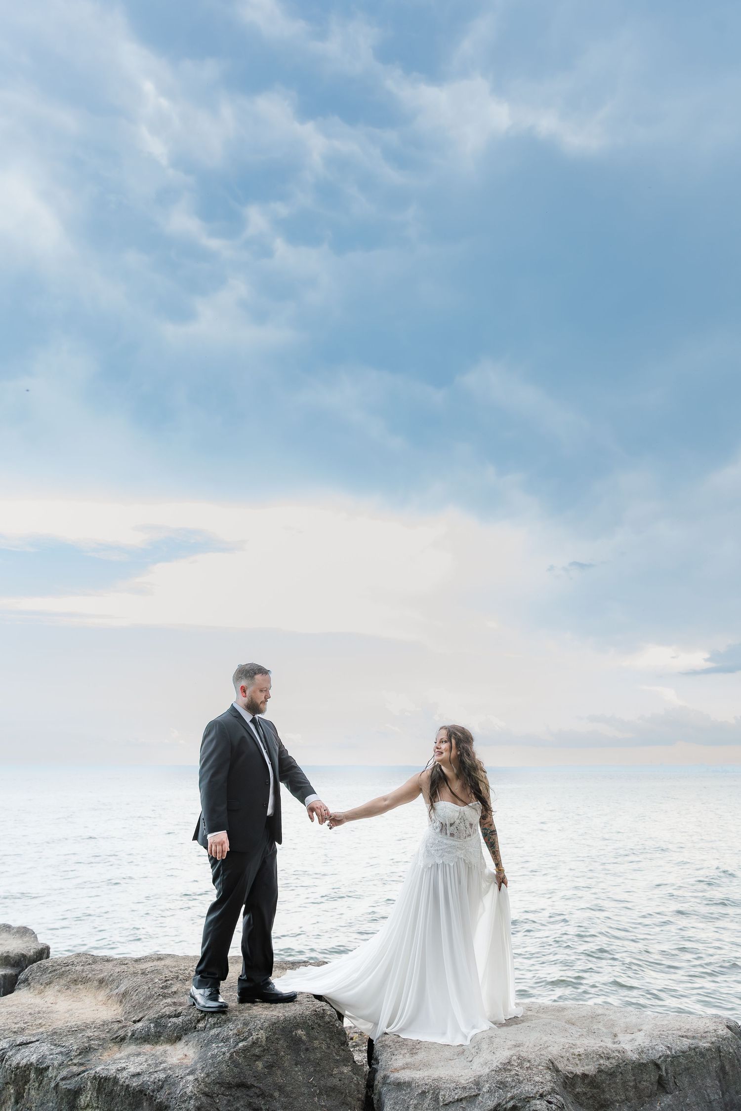 Bride leading the groom across rocks in front of lake ontario for natural wedding photos in burlington, ontario.