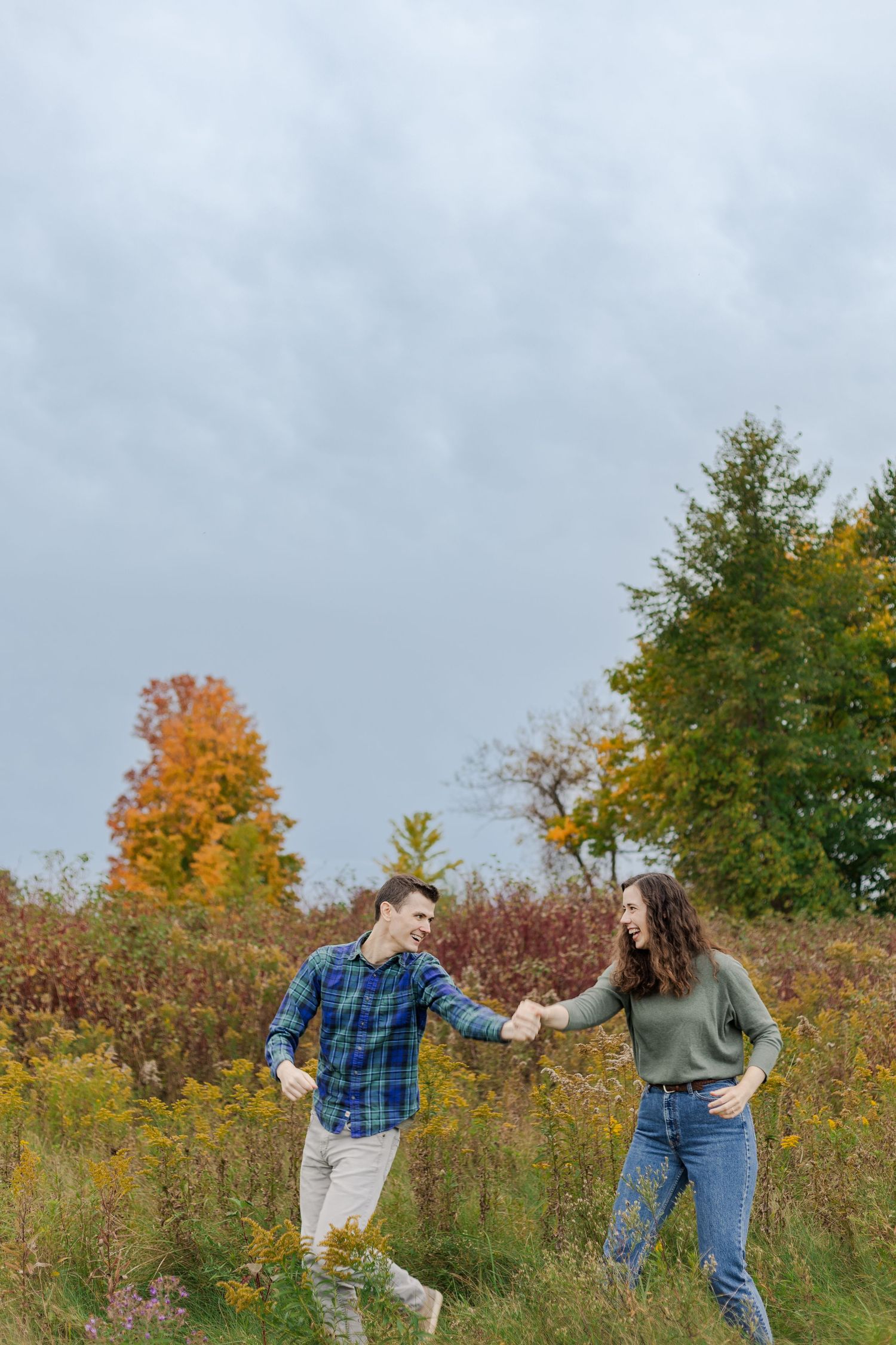 A couple dances and twirls through a fall meadow with colorful trees in background at Guelph Lake.